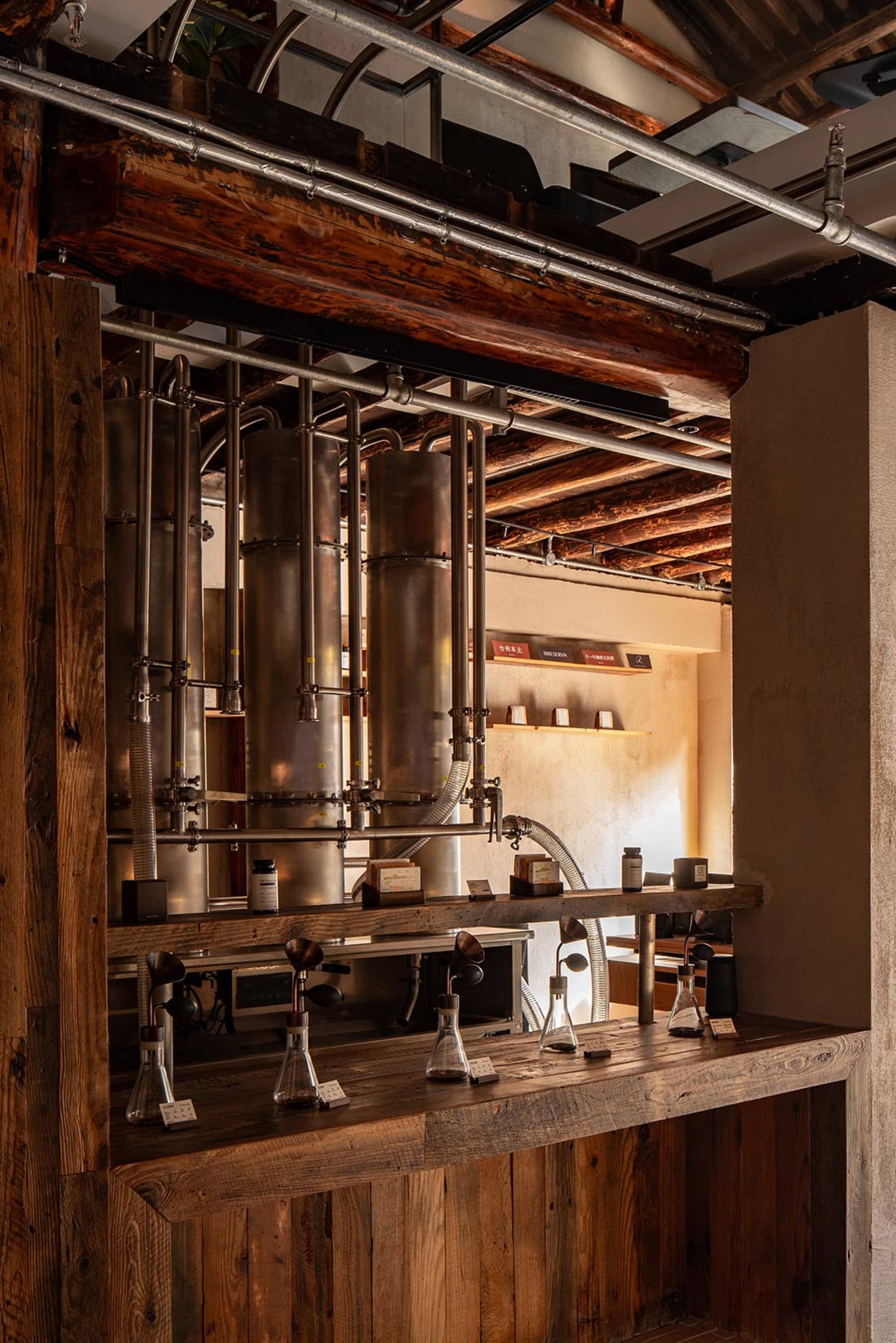 Interior view of an industrial-style distillery bar featuring large copper distillation columns, heavy reclaimed wood beams and counters, exposed metal pipes, and laboratory glassware under warm lighting.