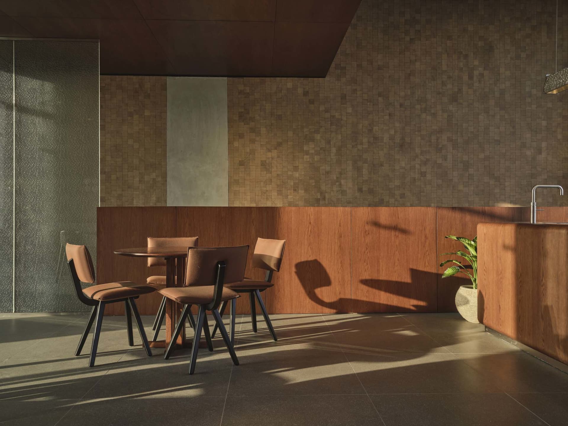 Modern minimalist dining area featuring a round walnut table, four terracotta upholstered chairs with black metal frames, wood-paneled walls, and dramatic natural light creating long shadows on a dark tiled floor.