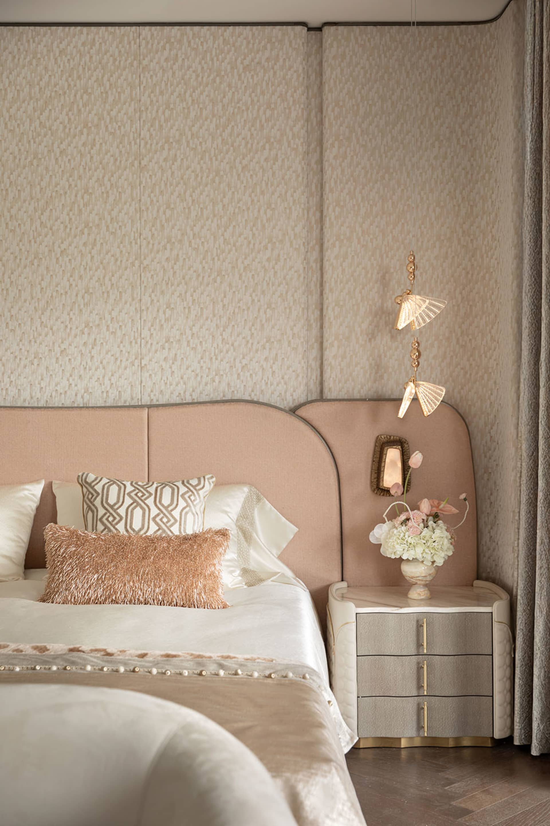 Modern luxury bedroom with blush pink curved upholstered headboard, textured beige wallpaper, white satin bedding, rose gold fur pillow, and elegant gold pendant light beside a marble nightstand with fresh flowers.