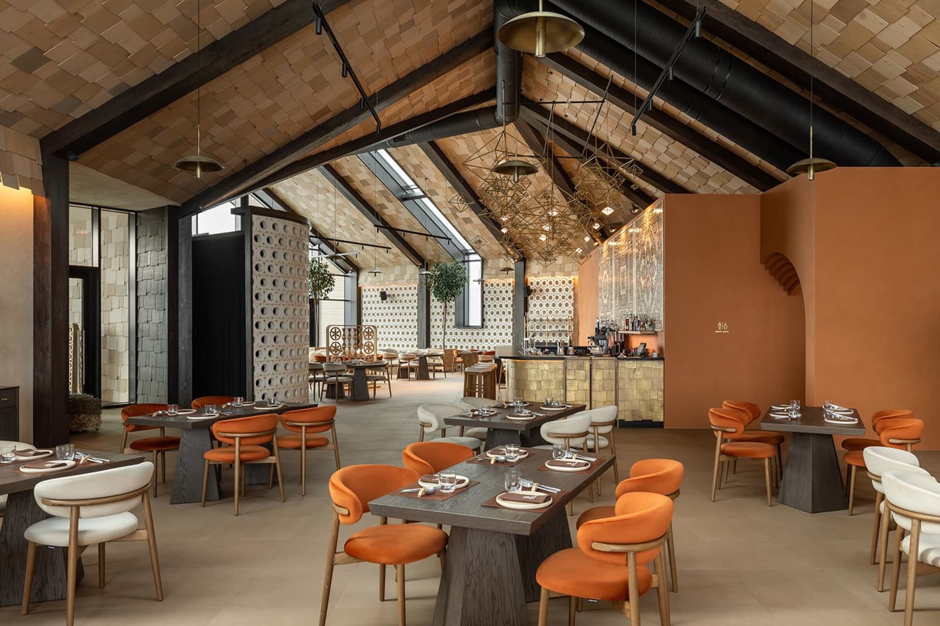 Modern restaurant interior featuring terracotta plaster walls, dark wood tables, orange and cream velvet chairs, and a dramatic vaulted ceiling with exposed wooden beams and geometric brass chandeliers.