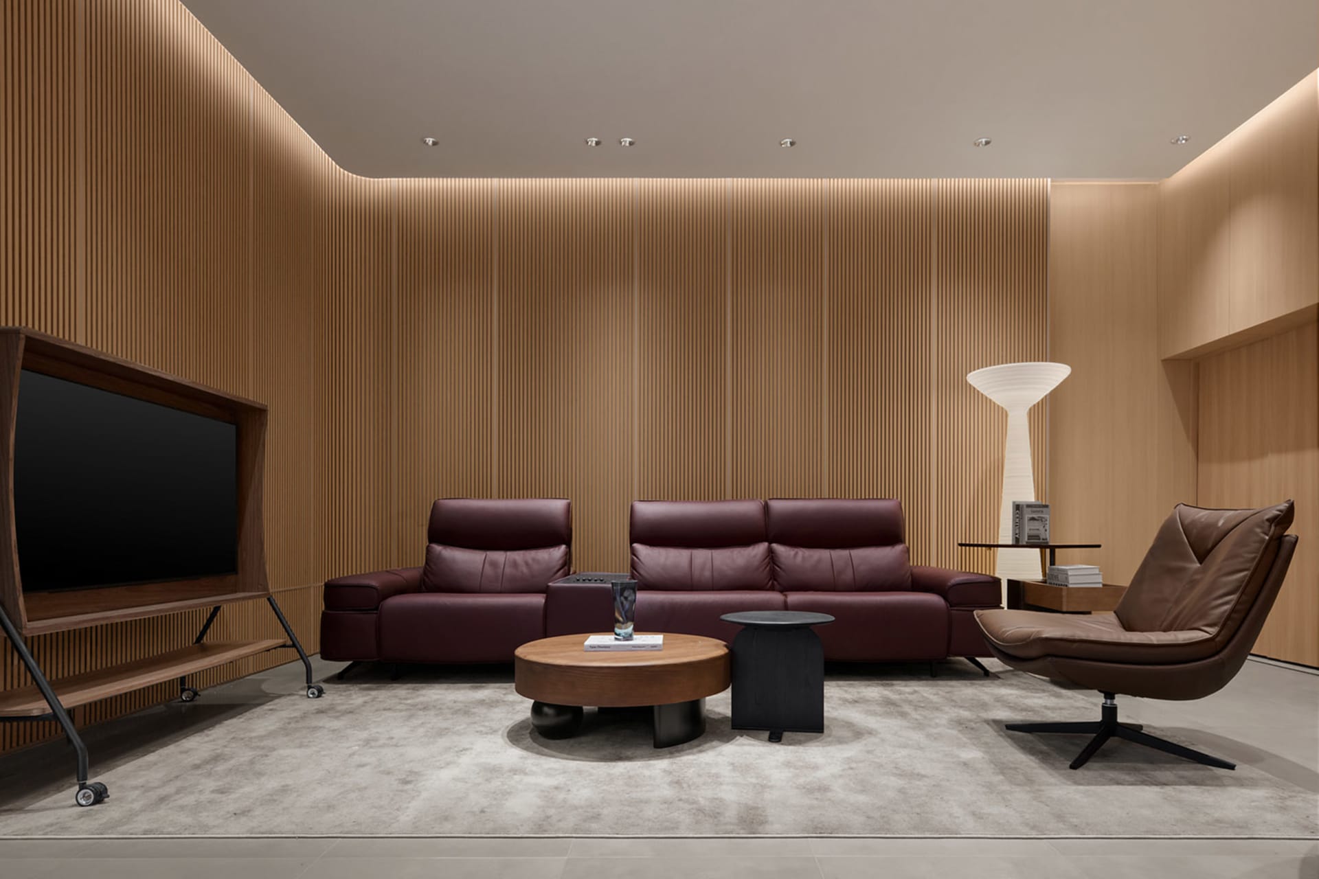 Modern minimalist living room featuring a burgundy leather sofa, matching loveseat, brown leather swivel chair, round wooden coffee table, vertical wood slat walls, and a white floor lamp on a light grey rug.
