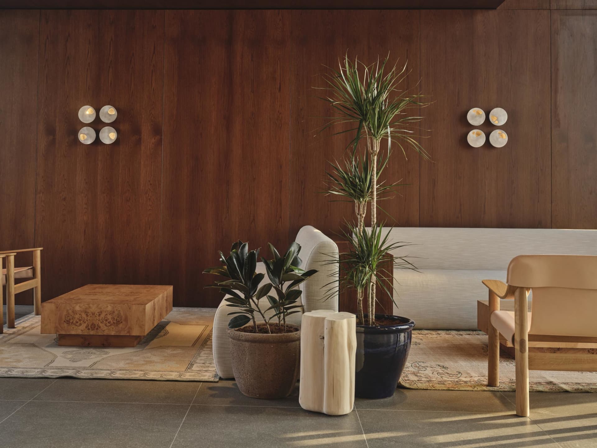 Modern living room featuring dark walnut wood panel walls, a large burl wood cube coffee table, light beige sofa, wooden armchair, potted plants, and layered area rugs on a tiled floor.