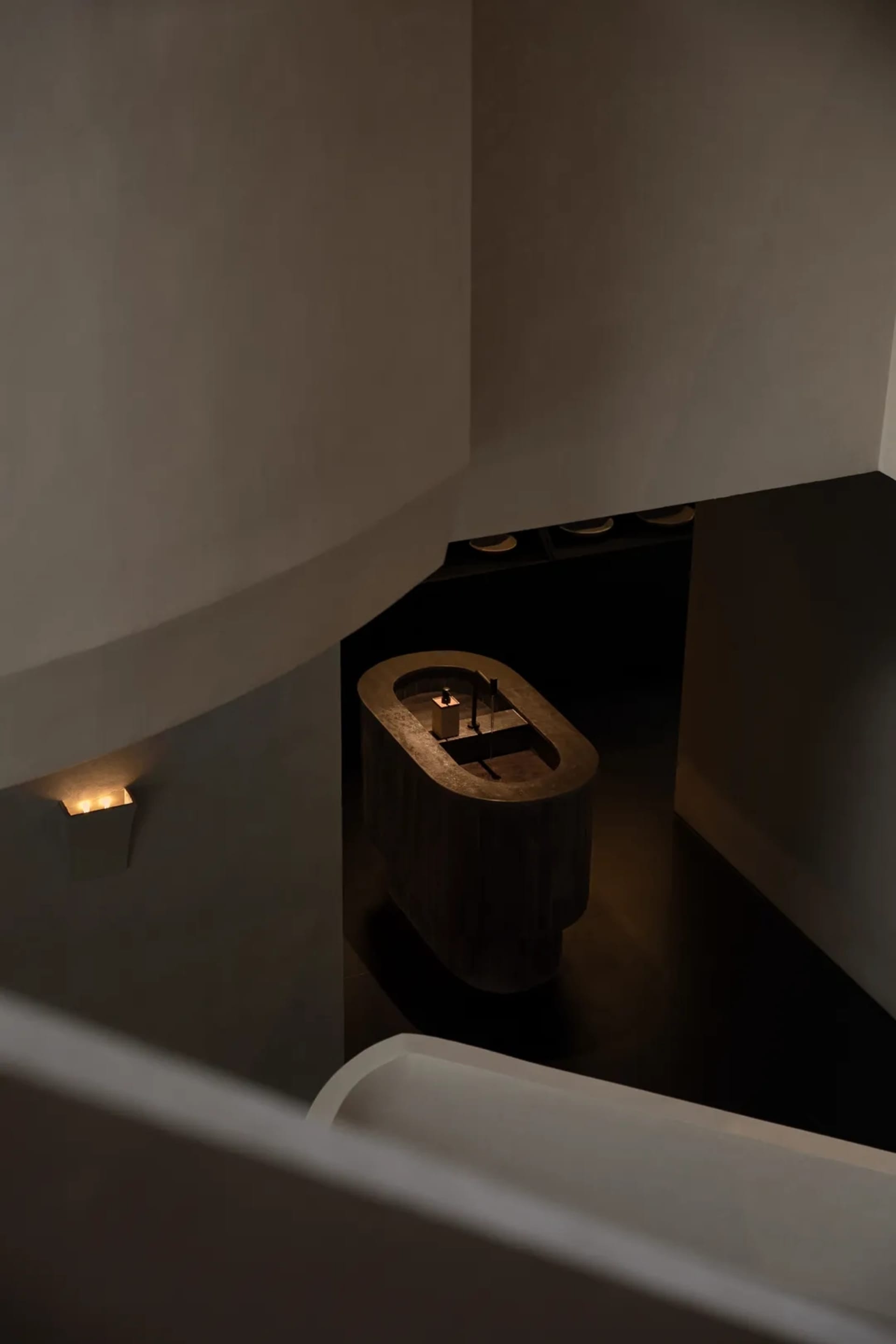 High-angle view of a minimalist dark bathroom featuring a freestanding concrete bathtub, recessed wall lighting, and dark flooring.