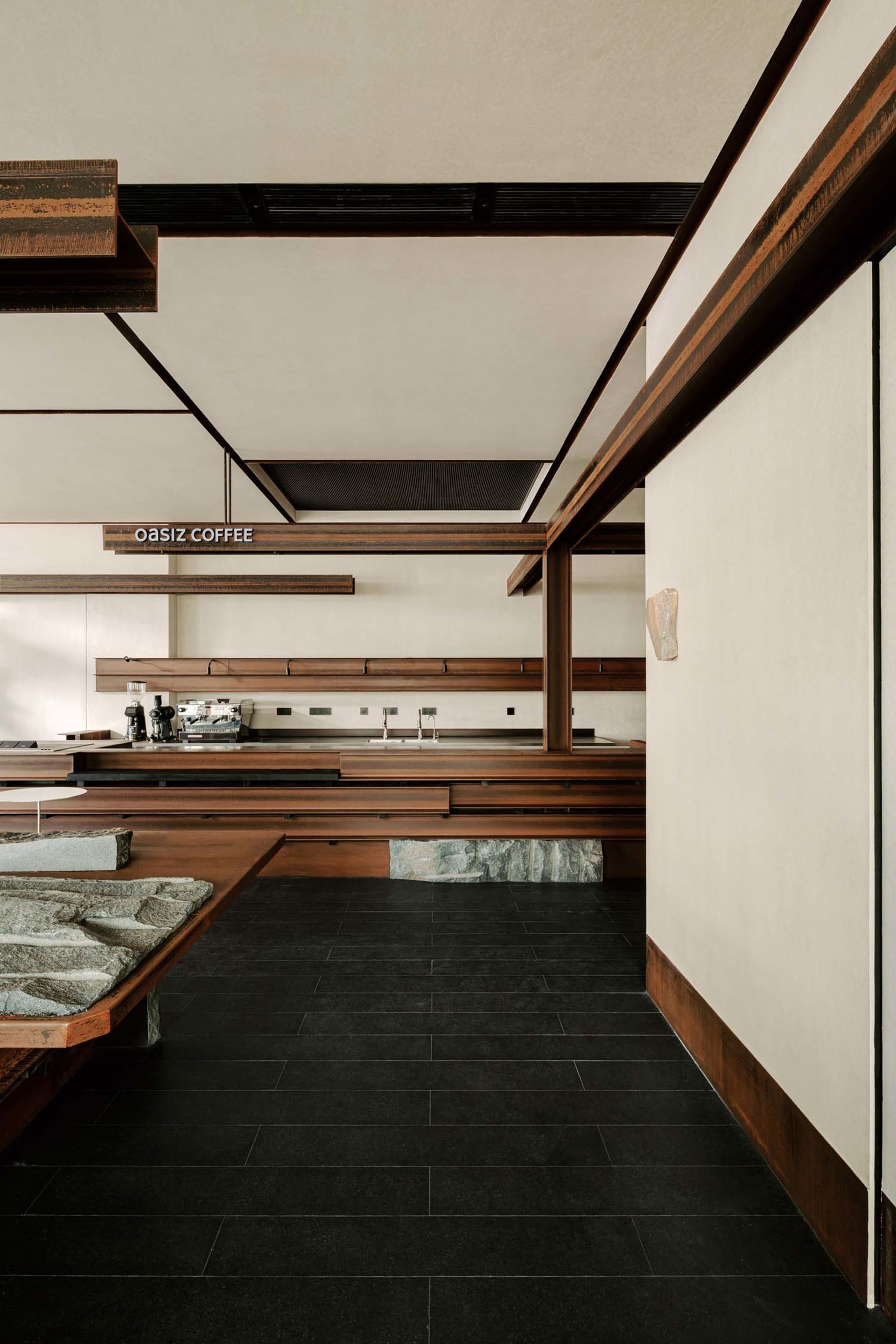 Modern minimalist cafe interior featuring dark walnut wood counters, stone elements, black tiled floor, and clean white walls with exposed structural beams.