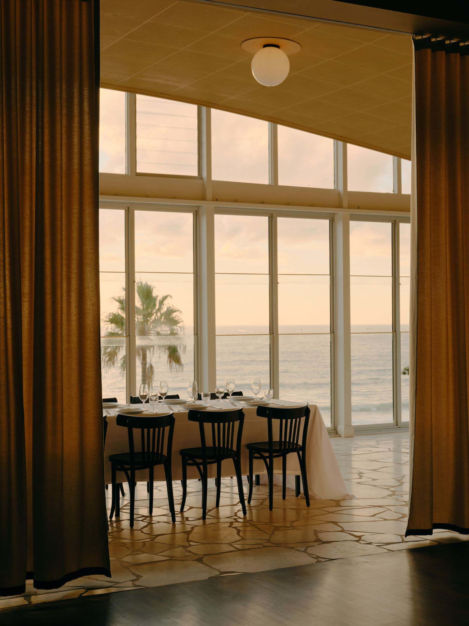 Elegant dining table set for guests in a room with large ocean-view windows, stone flooring, and heavy golden curtains at sunset.
