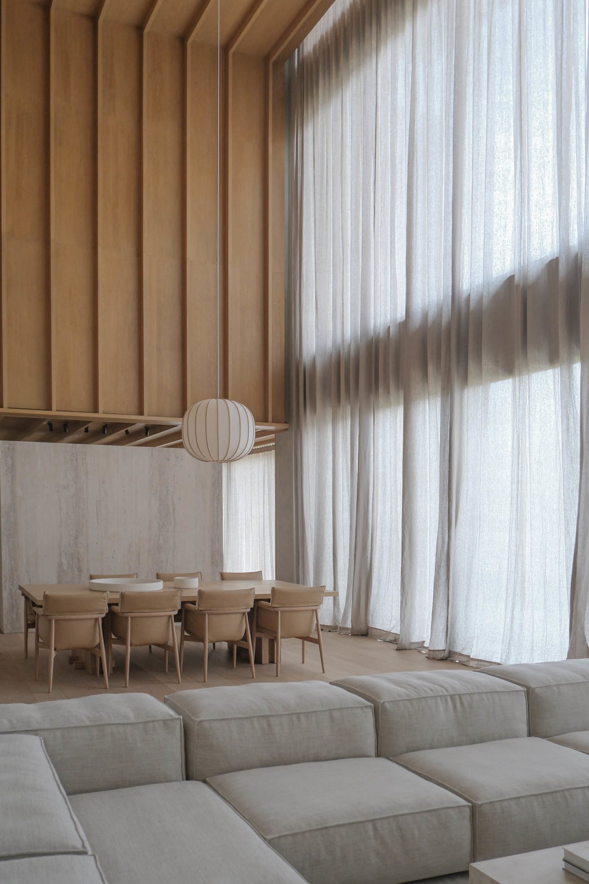 Modern minimalist dining area featuring a large oak table with beige upholstered chairs, a spherical ribbed pendant lamp, vertical oak wall paneling, and a textured concrete accent wall, viewed from a light-colored modular sofa in the foreground.