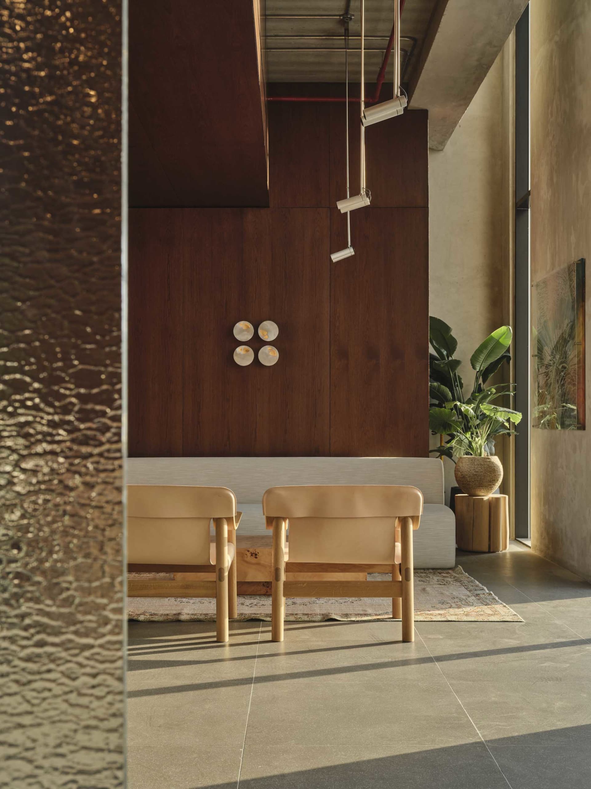 Modern minimalist living room featuring dark walnut wood wall paneling, two natural leather armchairs with light wood frames, a low wooden coffee table, and a large potted plant beside a concrete wall.