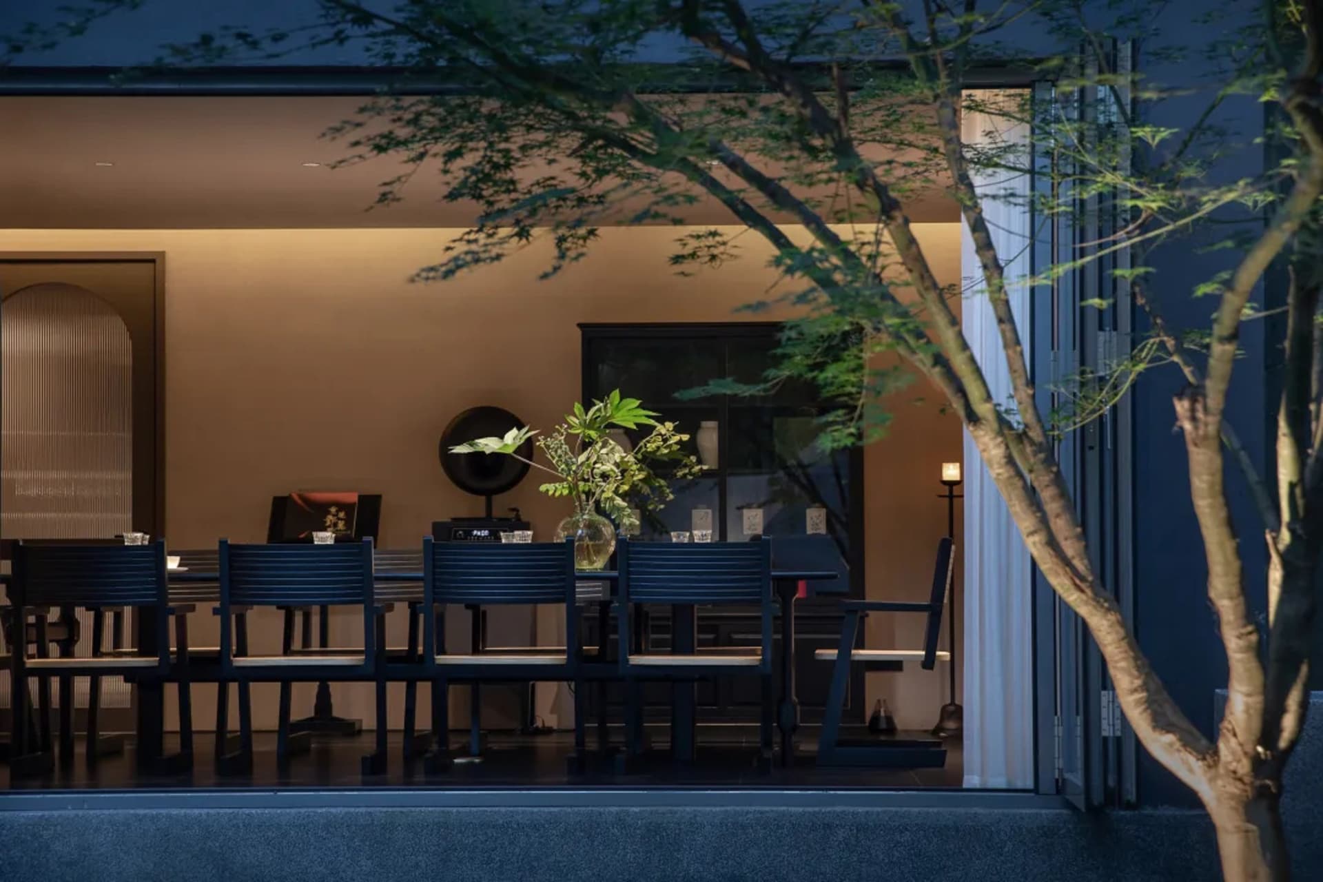 Modern minimalist dining room with dark blue wooden chairs and table, viewed through Japanese maple tree branches at dusk with warm interior lighting