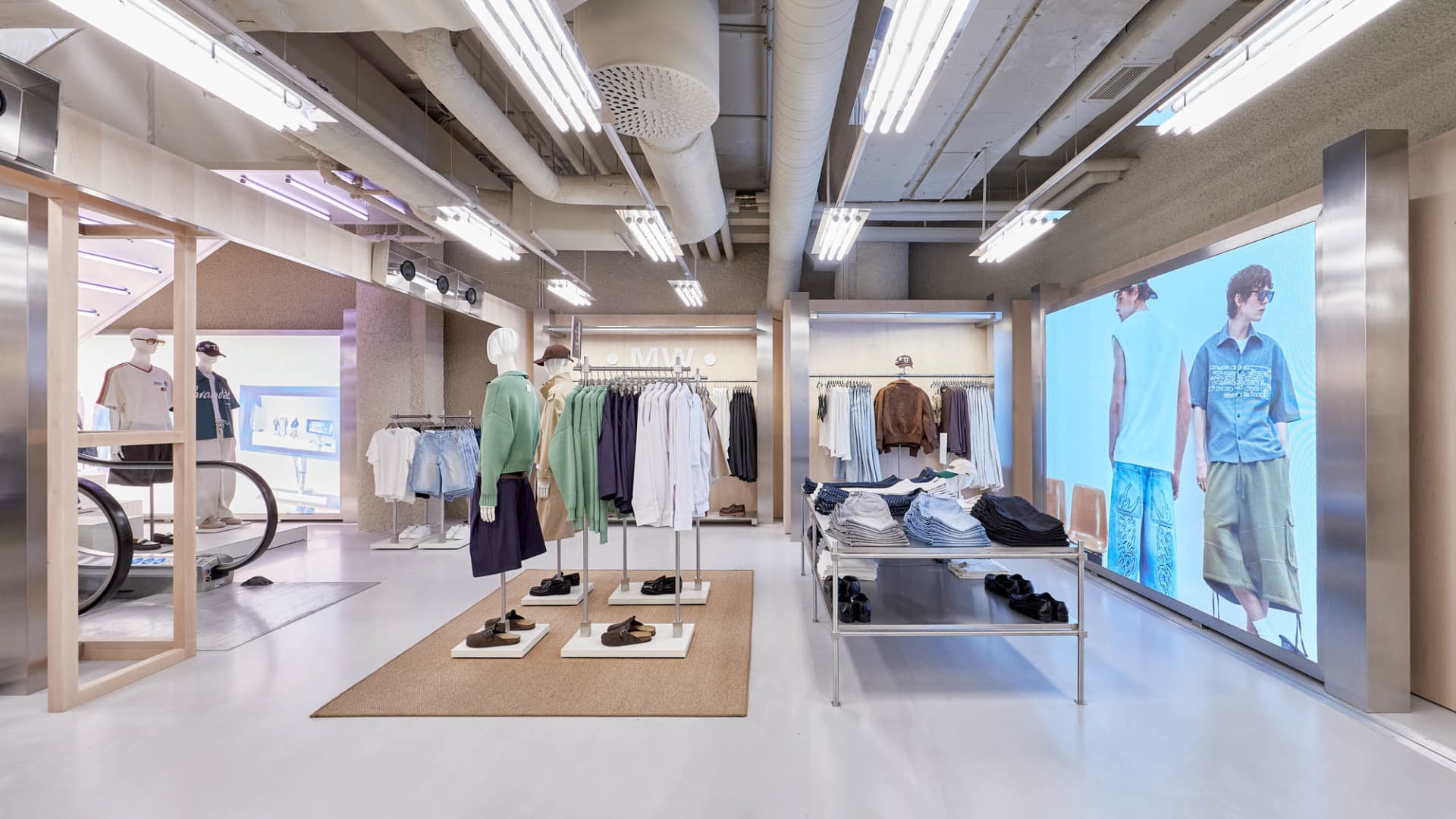 Bright, modern menswear retail interior with exposed concrete ceiling, linear fluorescent lighting, clothing racks, mannequins, and large digital screens displaying models.
