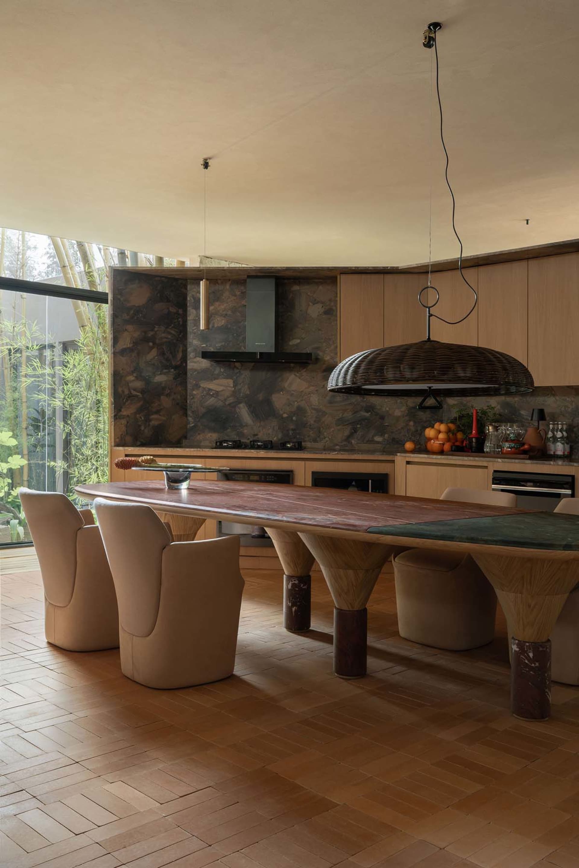 Modern dining room featuring a long curved walnut dining table with thick marble legs, beige upholstered chairs, a large woven pendant lamp, and a dramatic stone backsplash overlooking a bamboo garden.