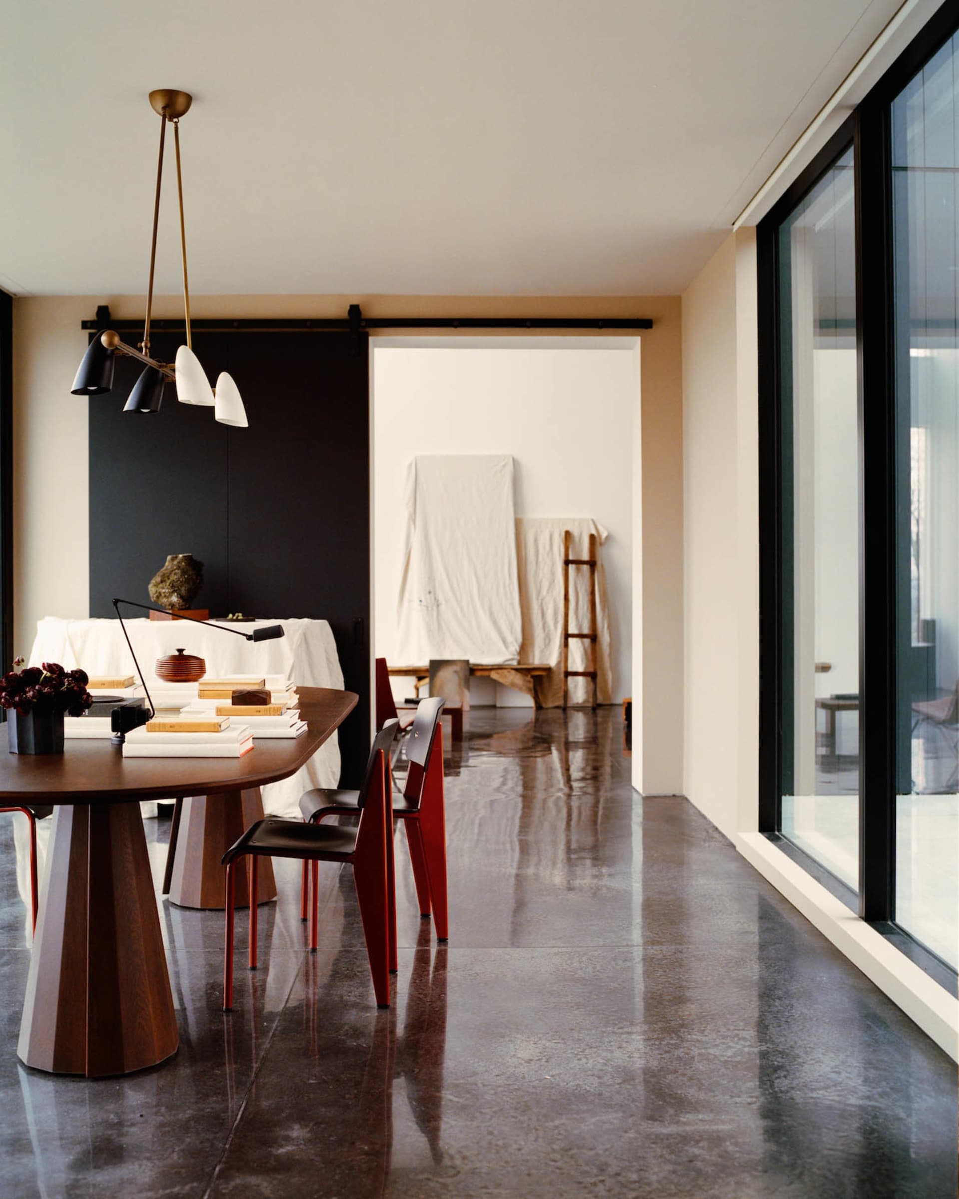 Modern minimalist dining room featuring a dark wood sculptural table, bright red dining chairs, polished concrete floor, black accent wall, and contemporary pendant light fixture.