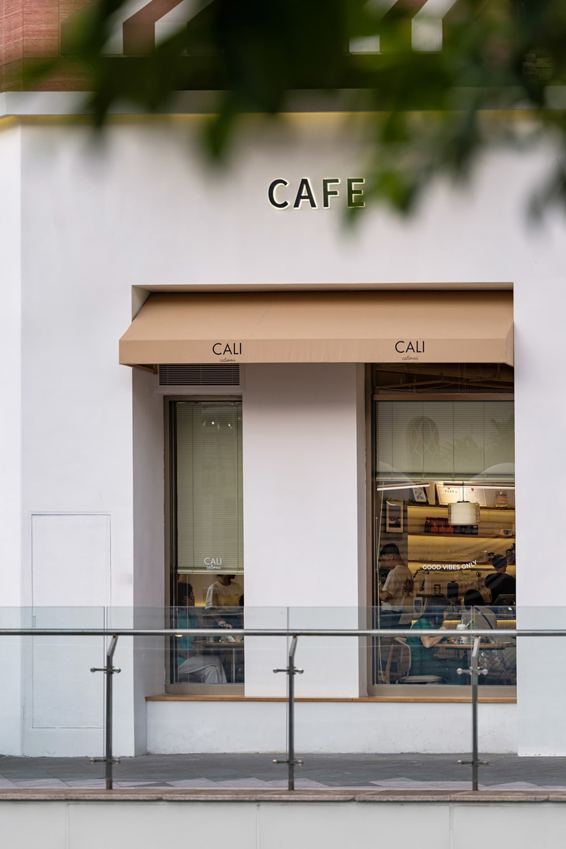 Street view of a modern white cafe named CALI with beige awnings, large glass windows showing customers inside, and a metal railing in the foreground.