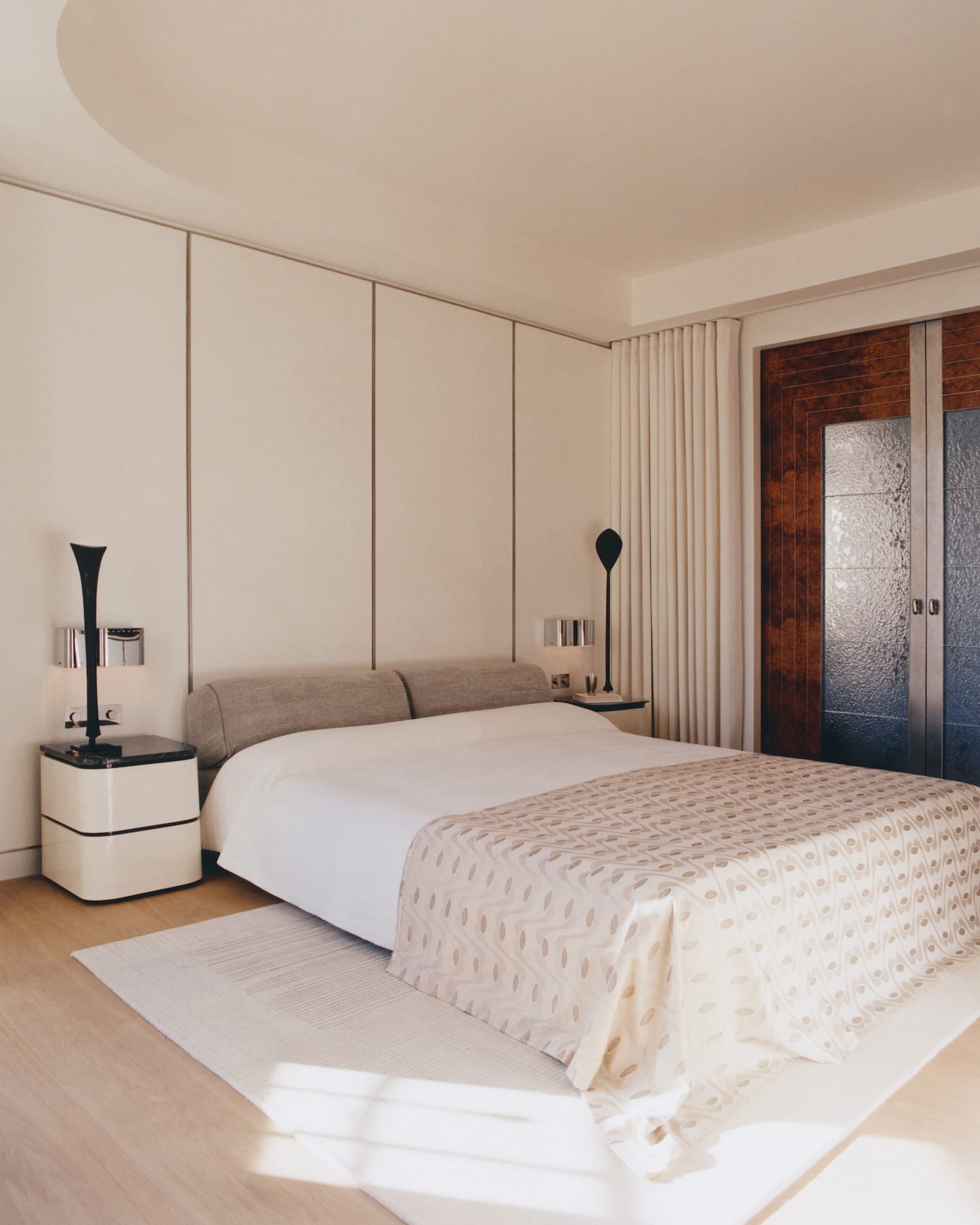 Modern minimalist bedroom with neutral beige walls, upholstered bed with patterned throw, white nightstand, black sculptural lamps, burl wood door, and bright natural sunlight on a light wood floor.