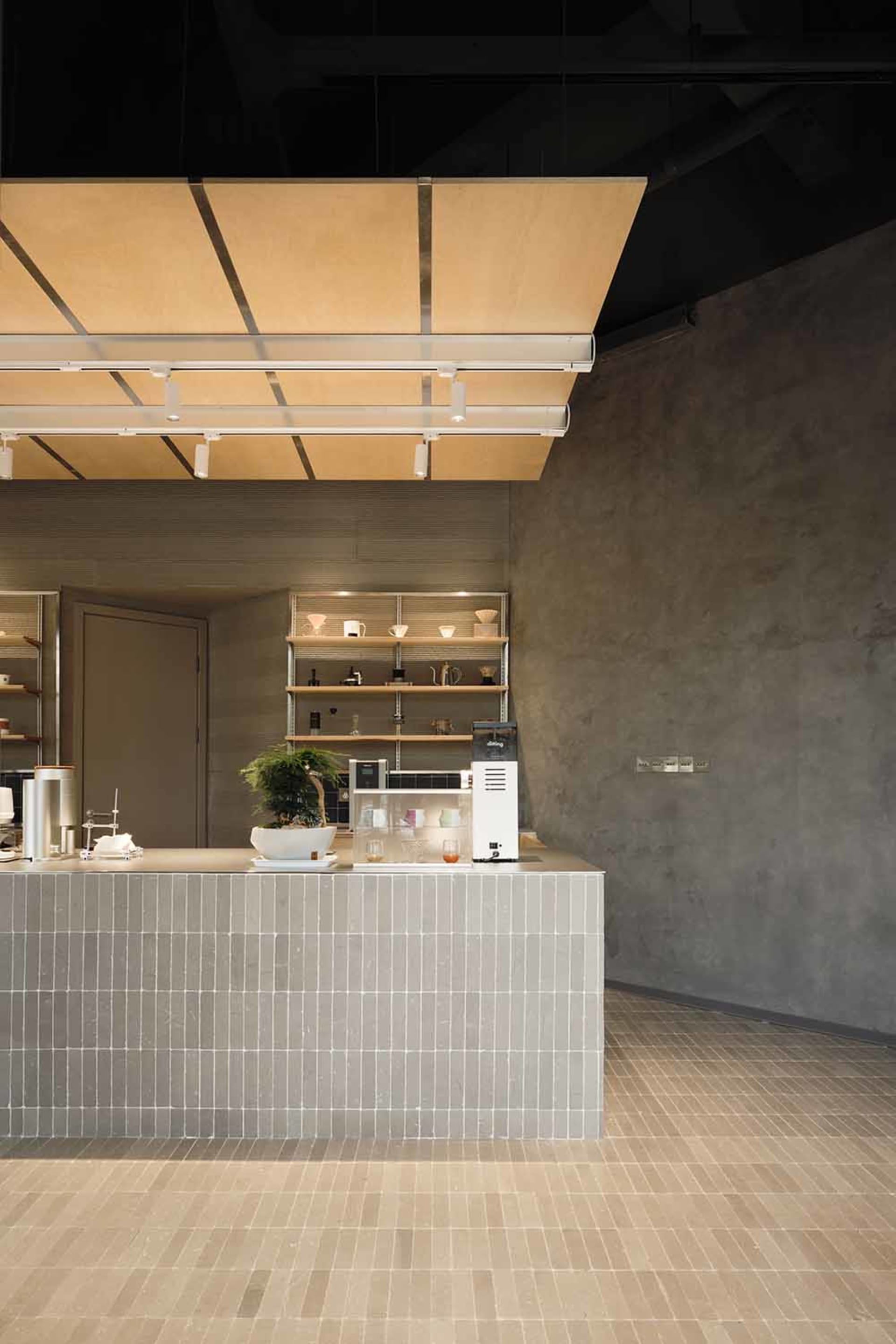 Modern minimalist cafe interior with a grey tiled concrete service counter, bonsai tree, coffee equipment, and a large floating wooden canopy with linear lighting.