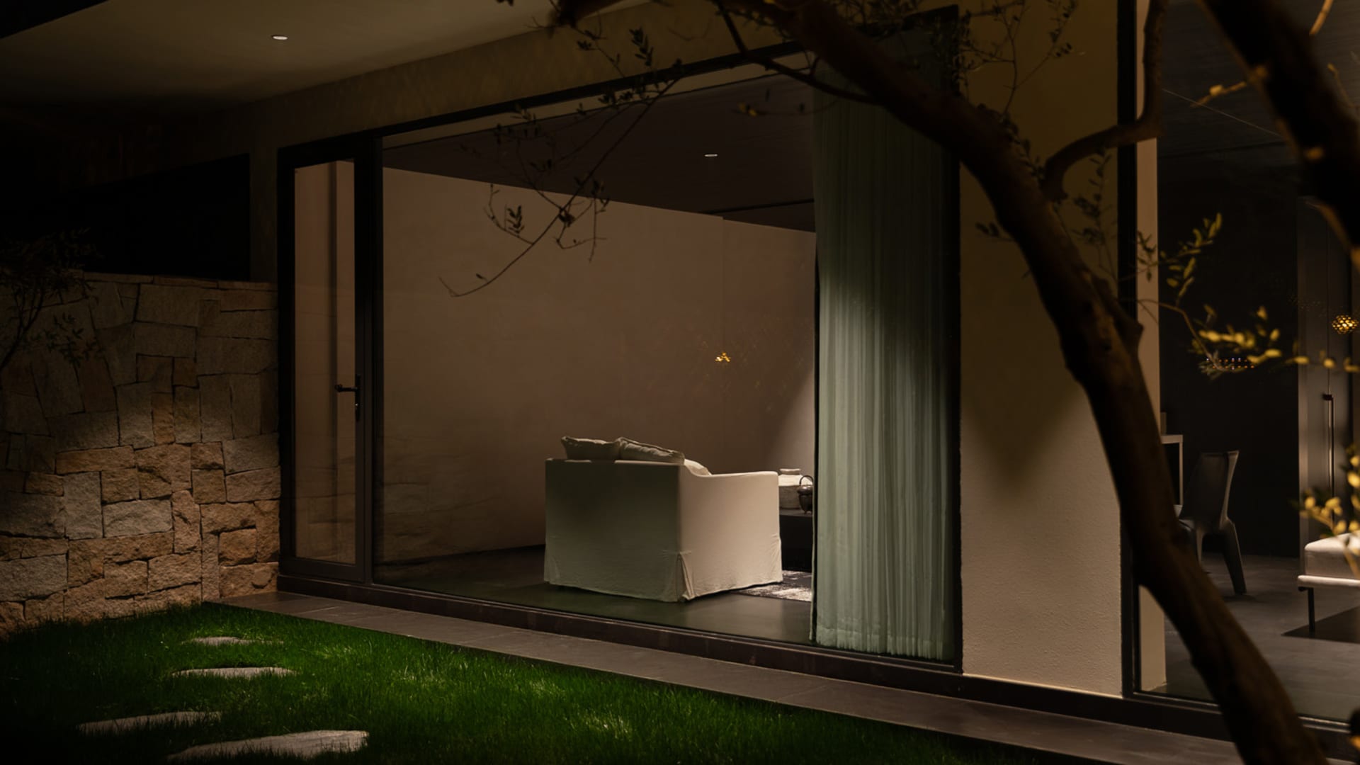 Exterior night view looking through large glass doors into a modern living room featuring a white sofa, textured stone wall, and warm ambient lighting.