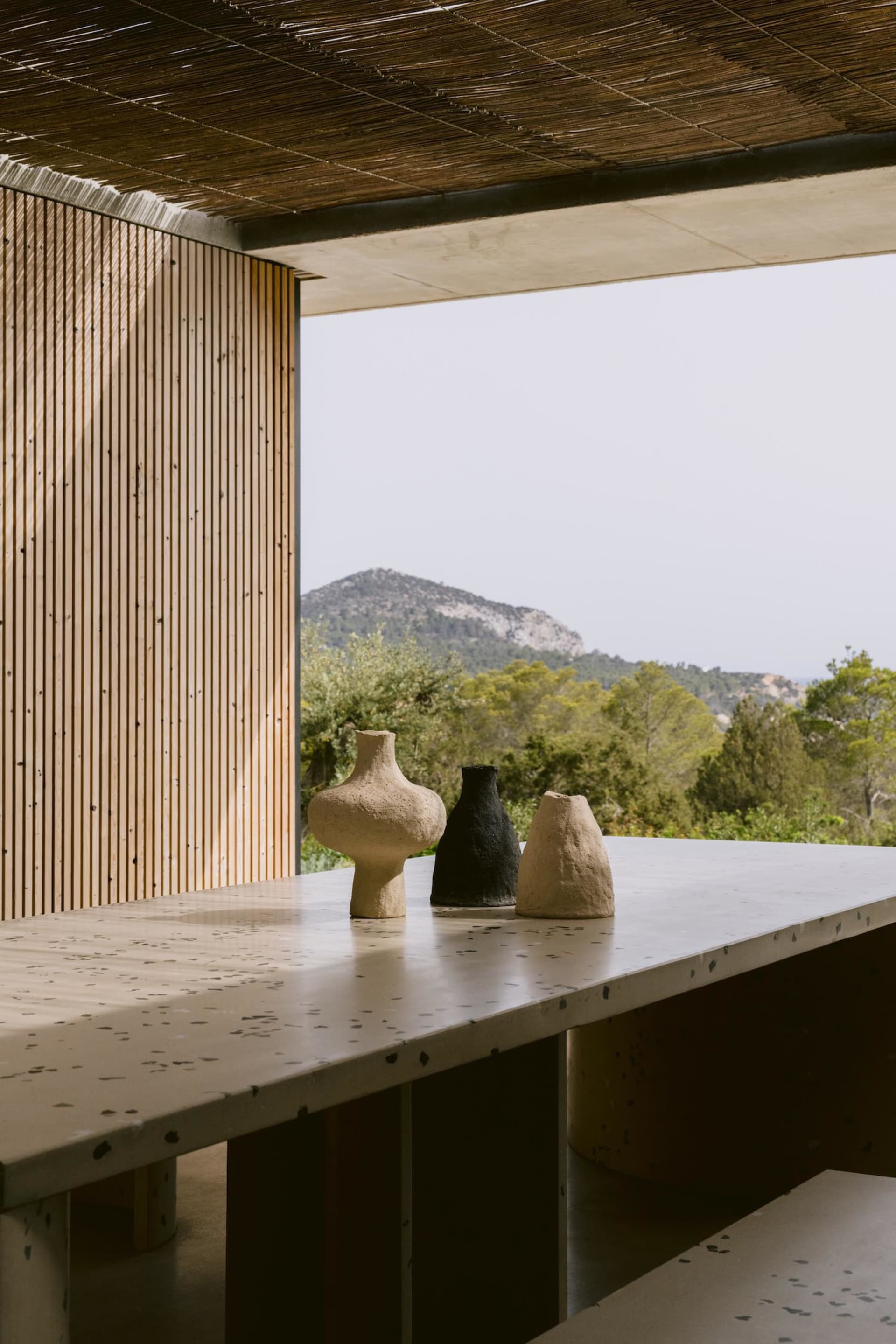 Speckled terrazzo counter with three rustic ceramic vases in beige, black and light terracotta, set against a wooden slat wall and panoramic mountain view under a thatched roof.