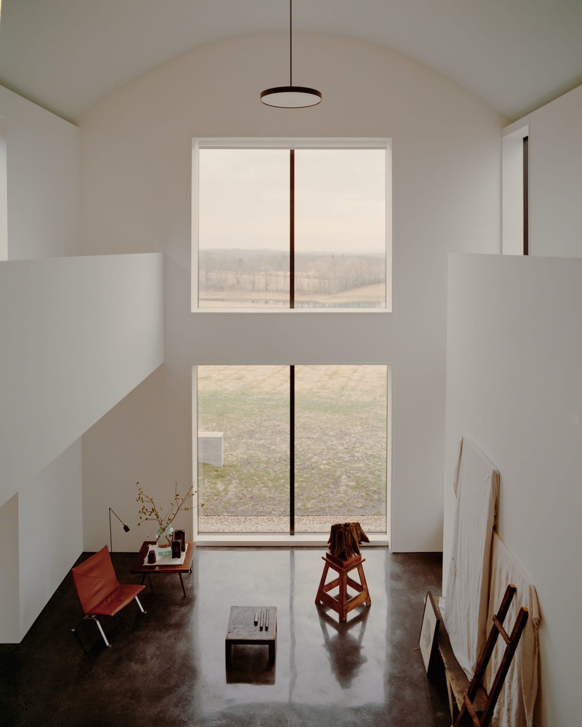 Minimalist double-height living room with polished concrete floor, orange leather chair, wooden stool, and large windows overlooking rural landscape