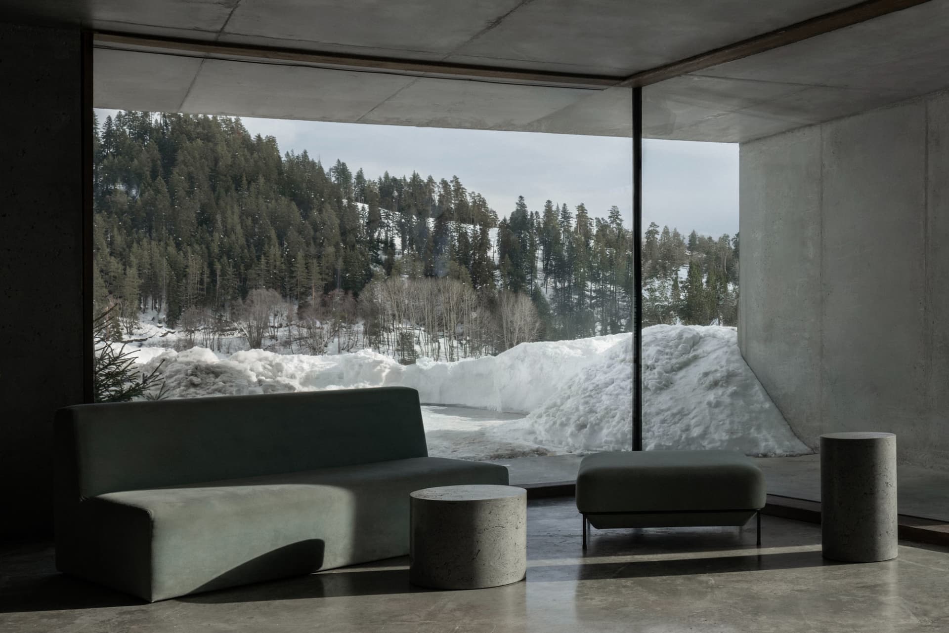 Modern minimalist living room with concrete walls and floor, featuring a curved green fabric sofa, matching ottoman, cylindrical concrete side tables, and a massive picture window overlooking a snowy forest and mountain.