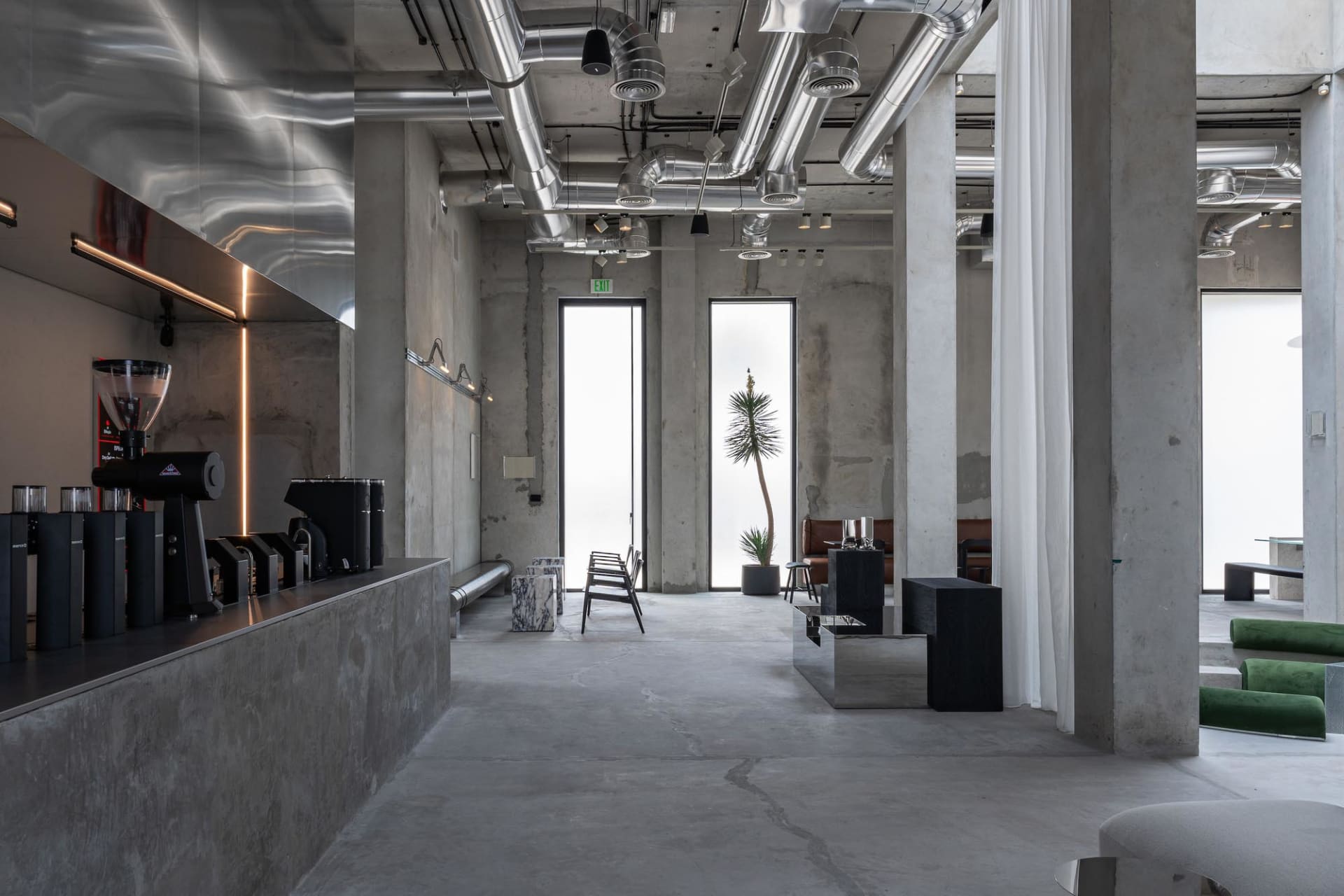 Modern industrial cafe interior featuring exposed concrete walls and floors, large stainless steel ventilation ducts, a coffee counter with espresso machines, and minimalist furniture under bright natural light from tall windows.