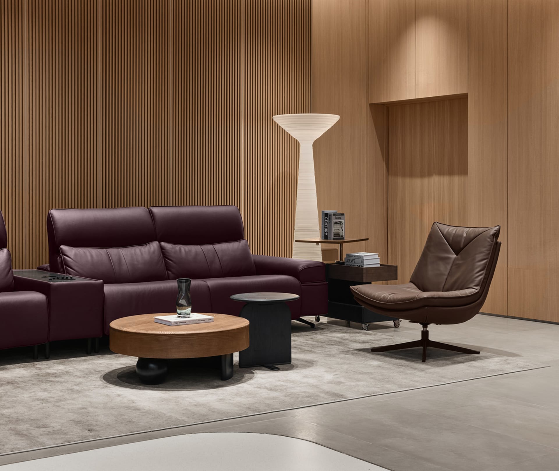 Modern living room with a burgundy leather sofa, brown leather swivel chair, round walnut coffee table, and vertical wood slat wall with a tall white floor lamp.