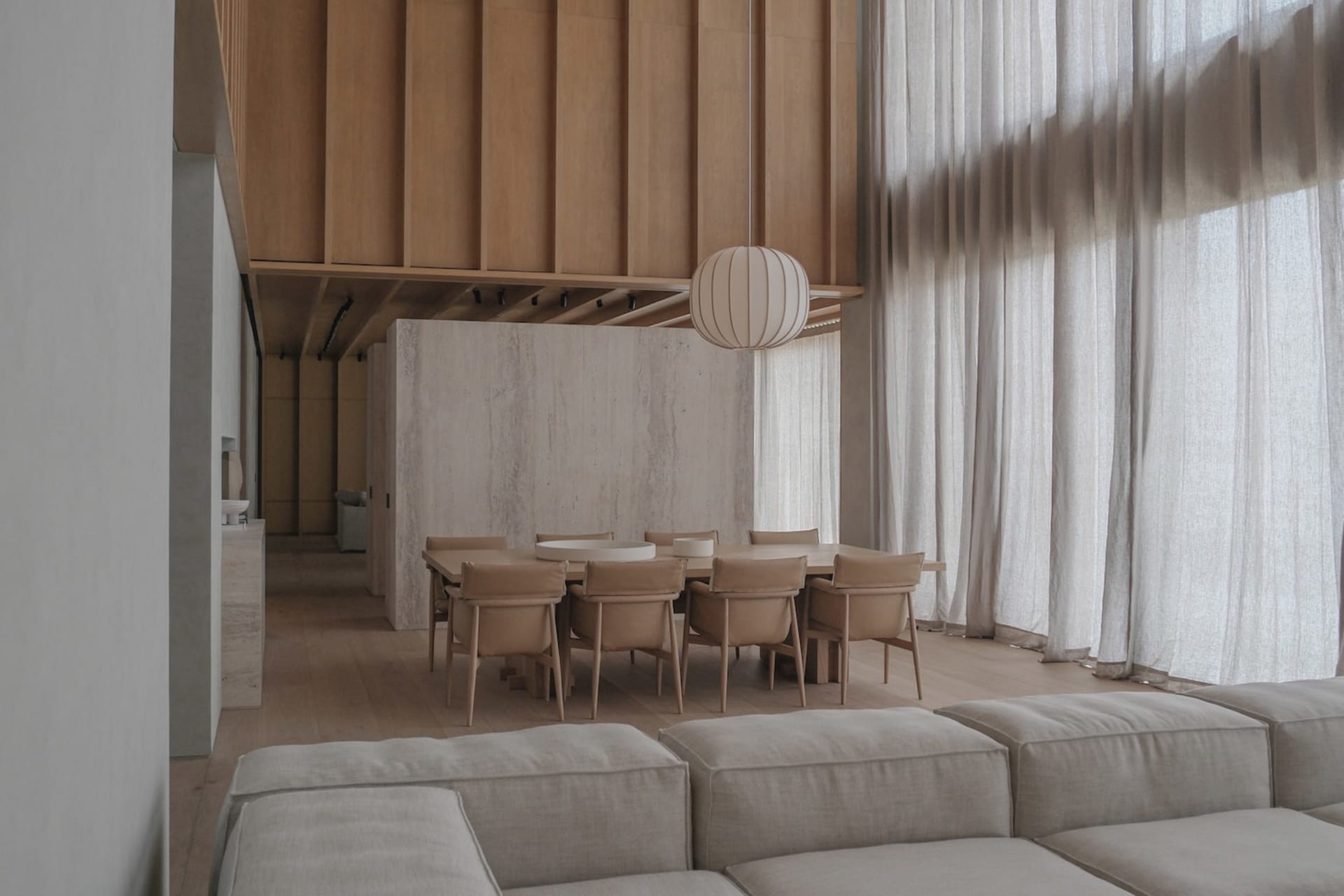 Modern minimalist dining area featuring a long oak table with beige upholstered chairs, a large paper lantern pendant light, concrete accent wall, and light oak flooring, viewed from a neutral-toned sofa.
