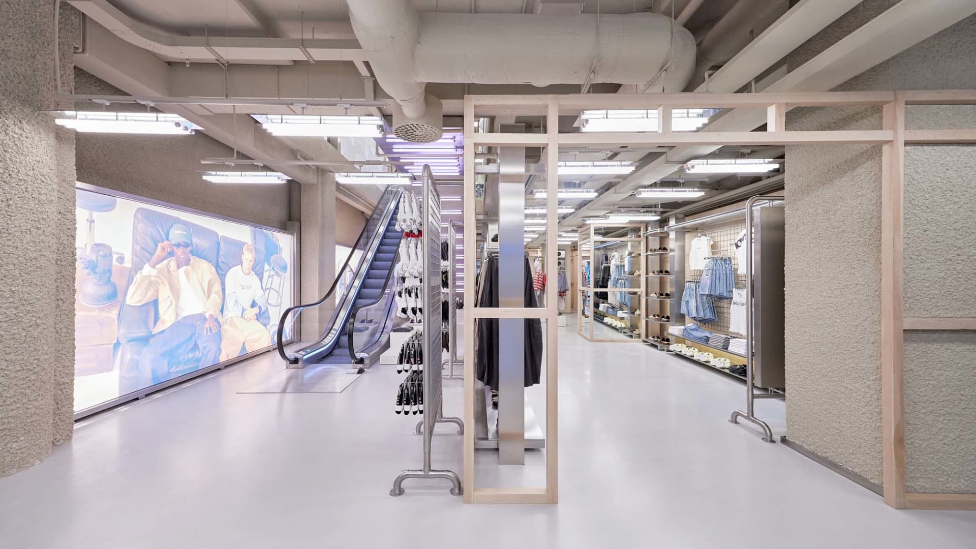 現代工業風格服裝店內部,擁有清水模牆面、木質模組化展示結構、服裝吊架、大型數位牆面顯示器以及手扶梯。