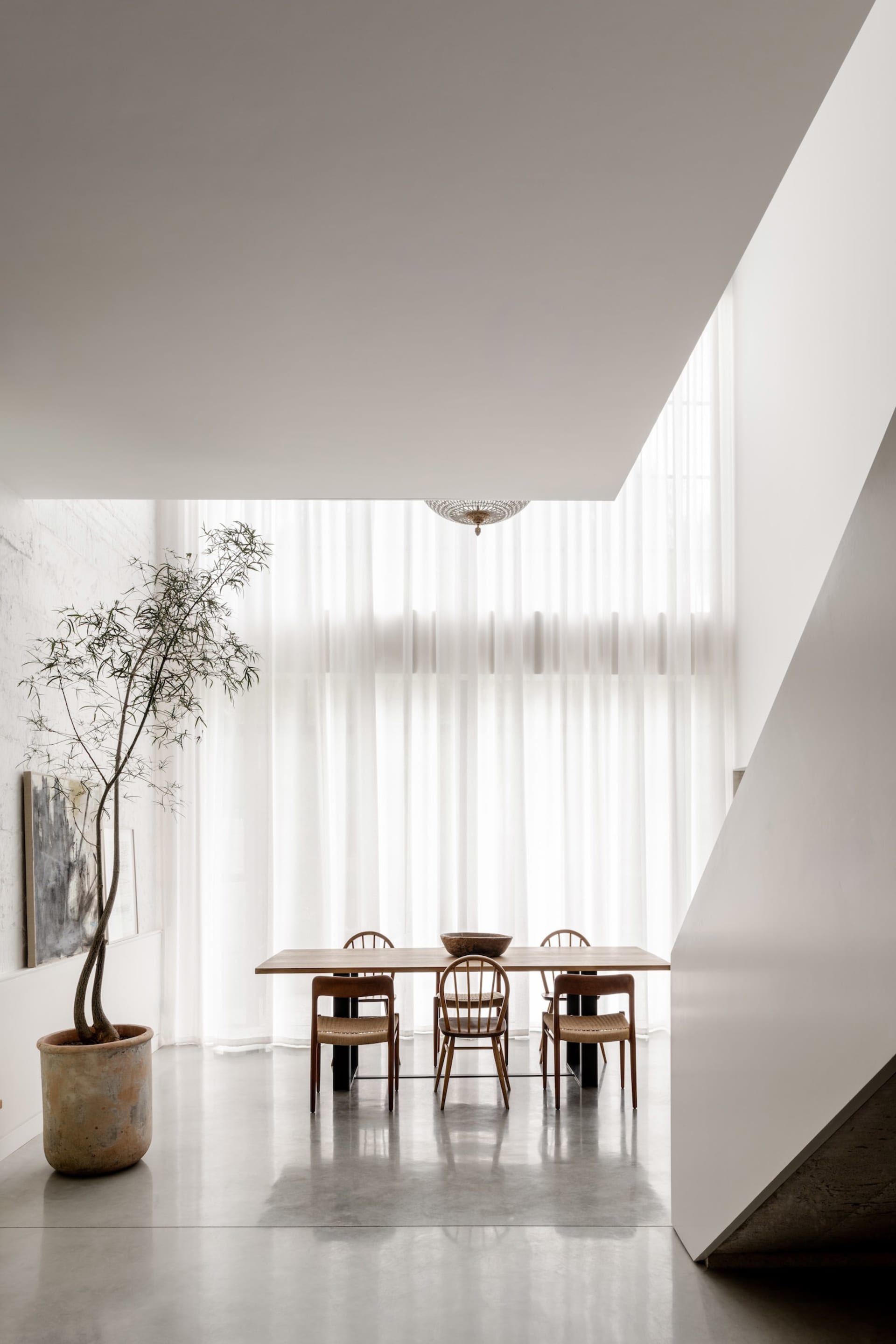 Minimalist dining room featuring a long oak wood table, four wooden chairs with woven seats, a large potted tree, sheer white curtains, and a woven pendant light.