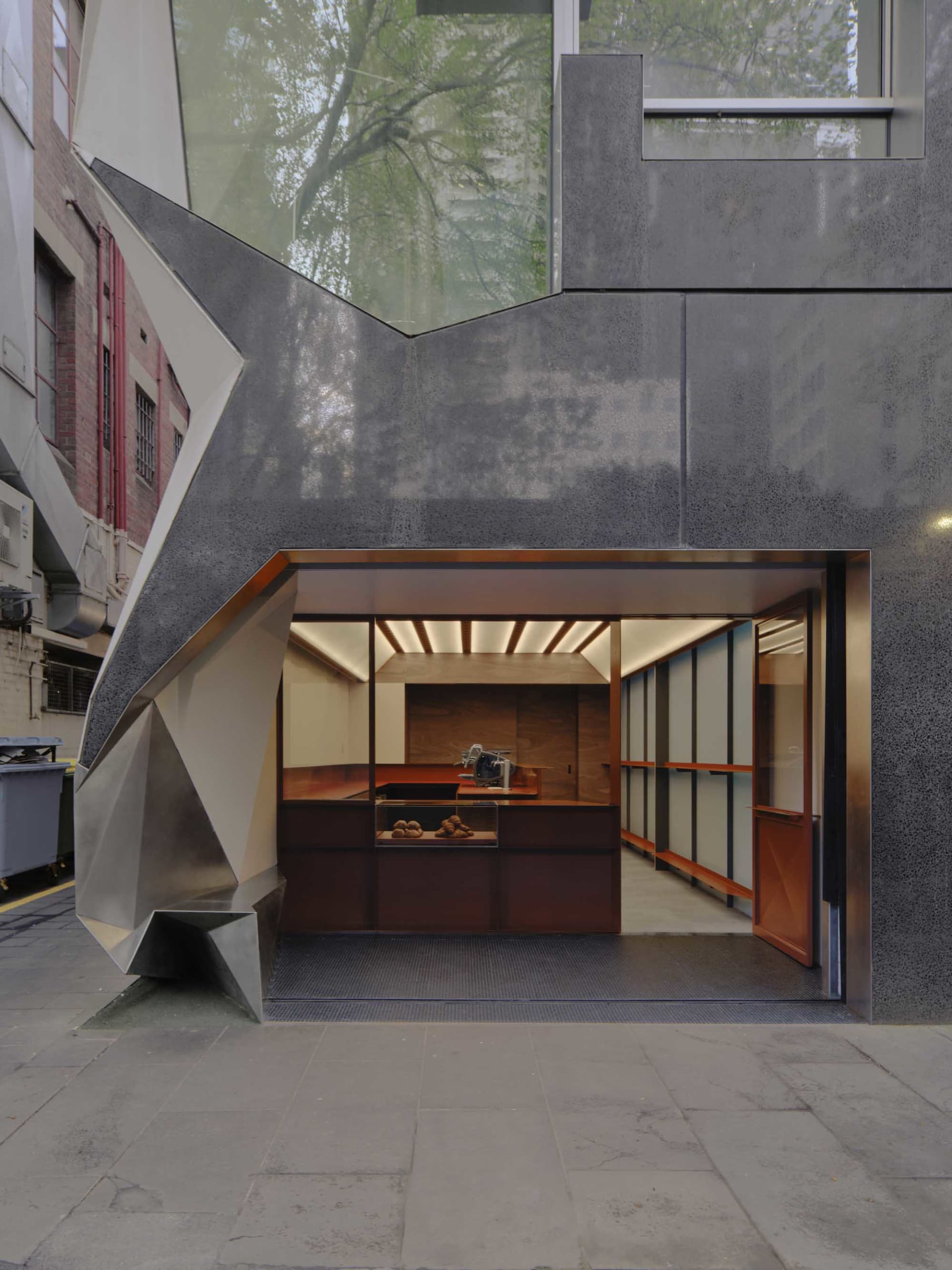 Modern bakery storefront featuring a dramatic angular facade made of weathered corten steel and concrete, with an open view into a wood-lined interior displaying fresh pastries.