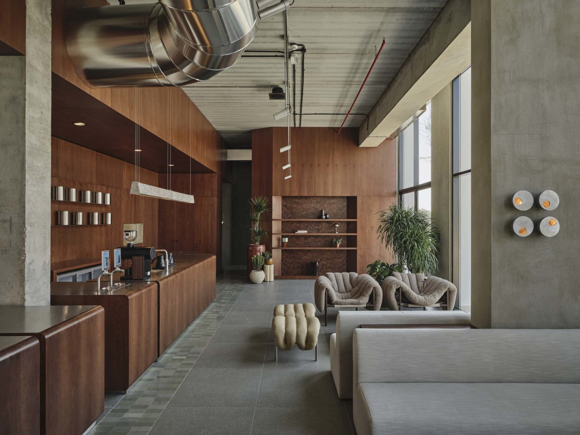 Modern industrial cafe lounge with extensive walnut wood cabinetry, concrete walls, large windows, boucle armchairs, and a prominent stainless steel ventilation duct suspended from the ceiling.
