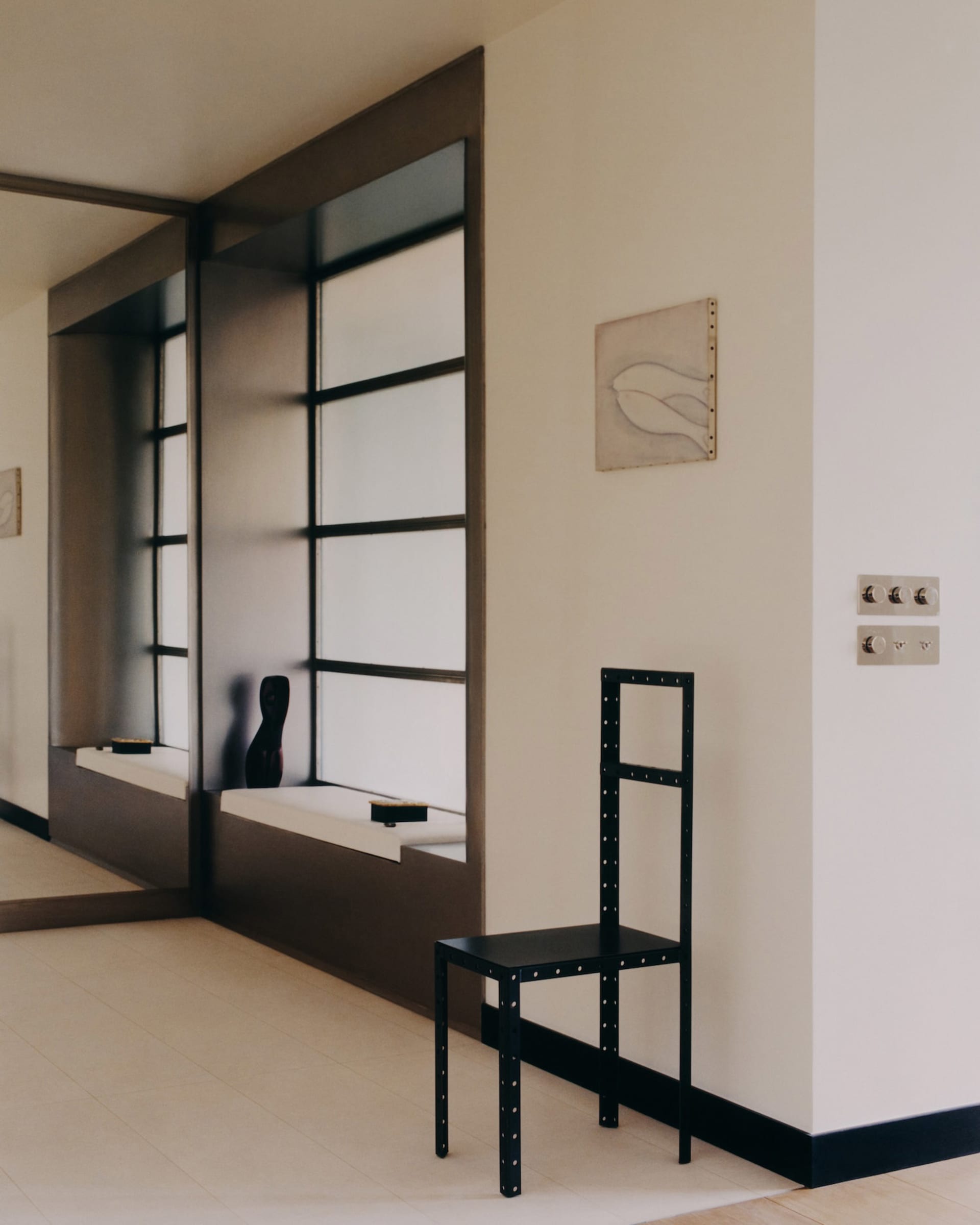 Modern minimalist entryway featuring a built-in dark wood window niche with white seat, a tall black metal chair with rivet details, and a framed abstract line drawing on a cream wall.