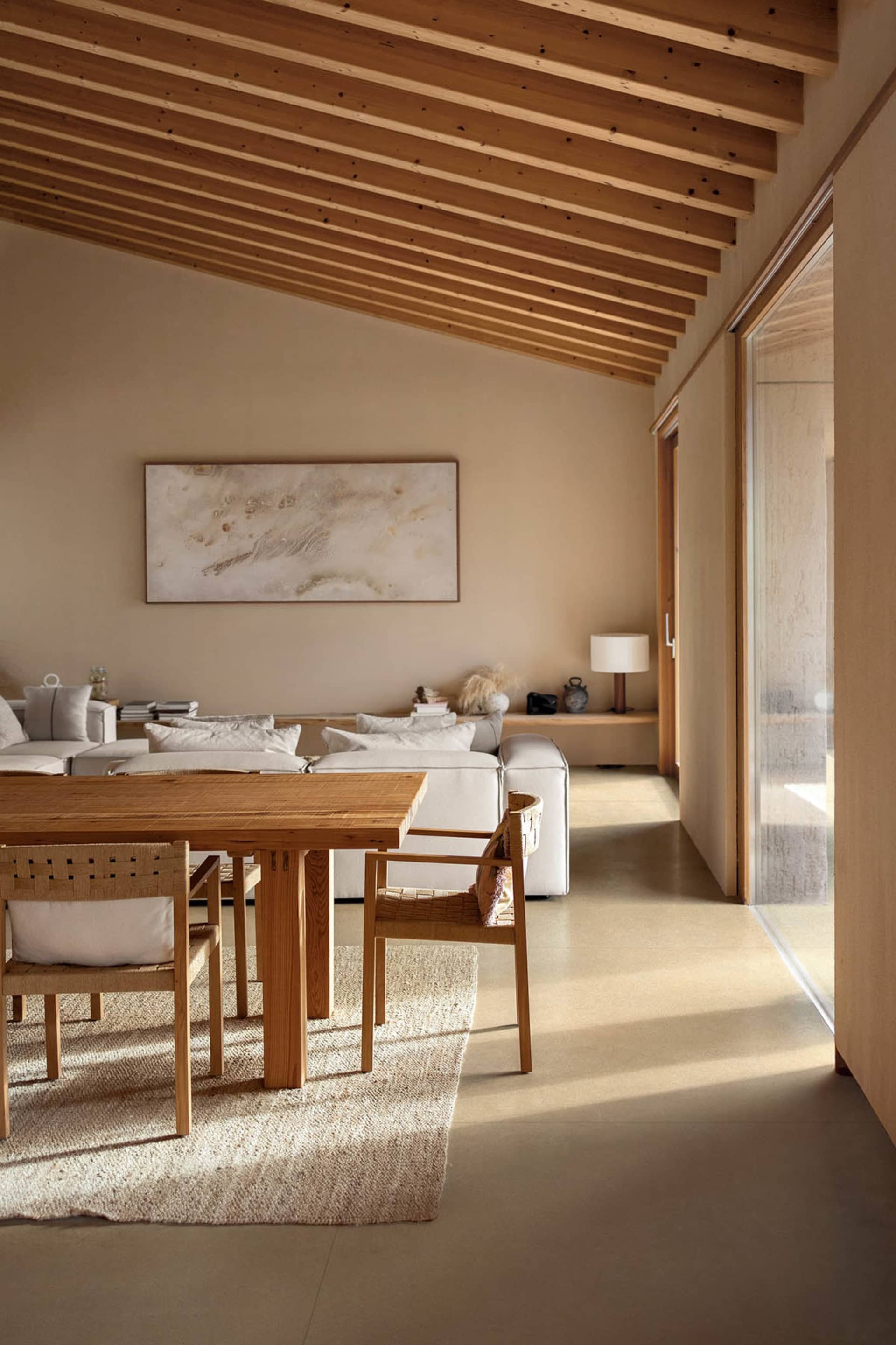 Modern minimalist dining area with solid oak table and woven rattan chairs on a textured rug, white sectional sofa in the background, exposed wooden beam ceiling and large abstract painting on the wall.