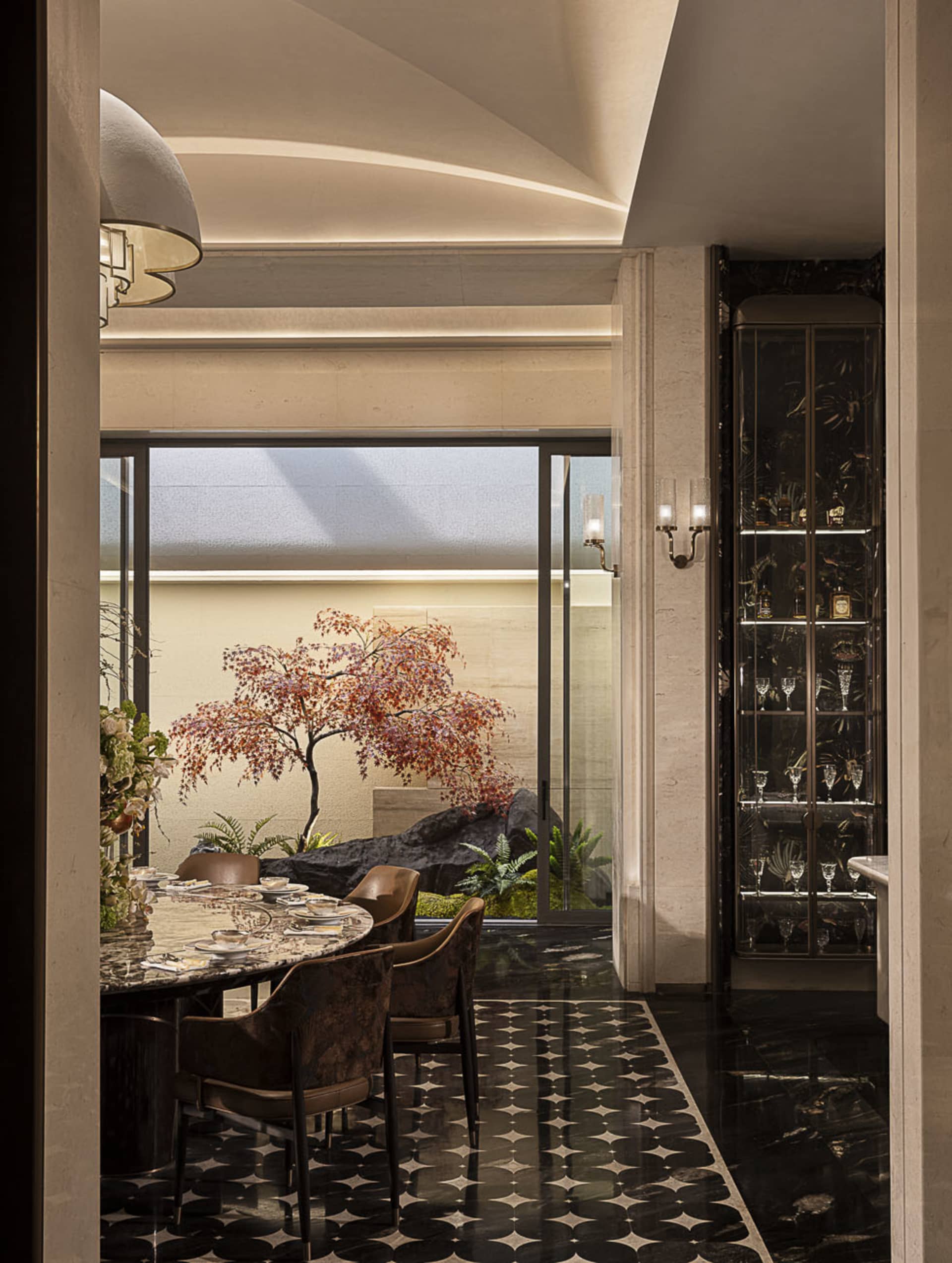 Modern luxury dining room with round marble table, velvet dining chairs, black and white patterned marble floor, and a Japanese maple tree in an indoor courtyard visible through large glass doors.