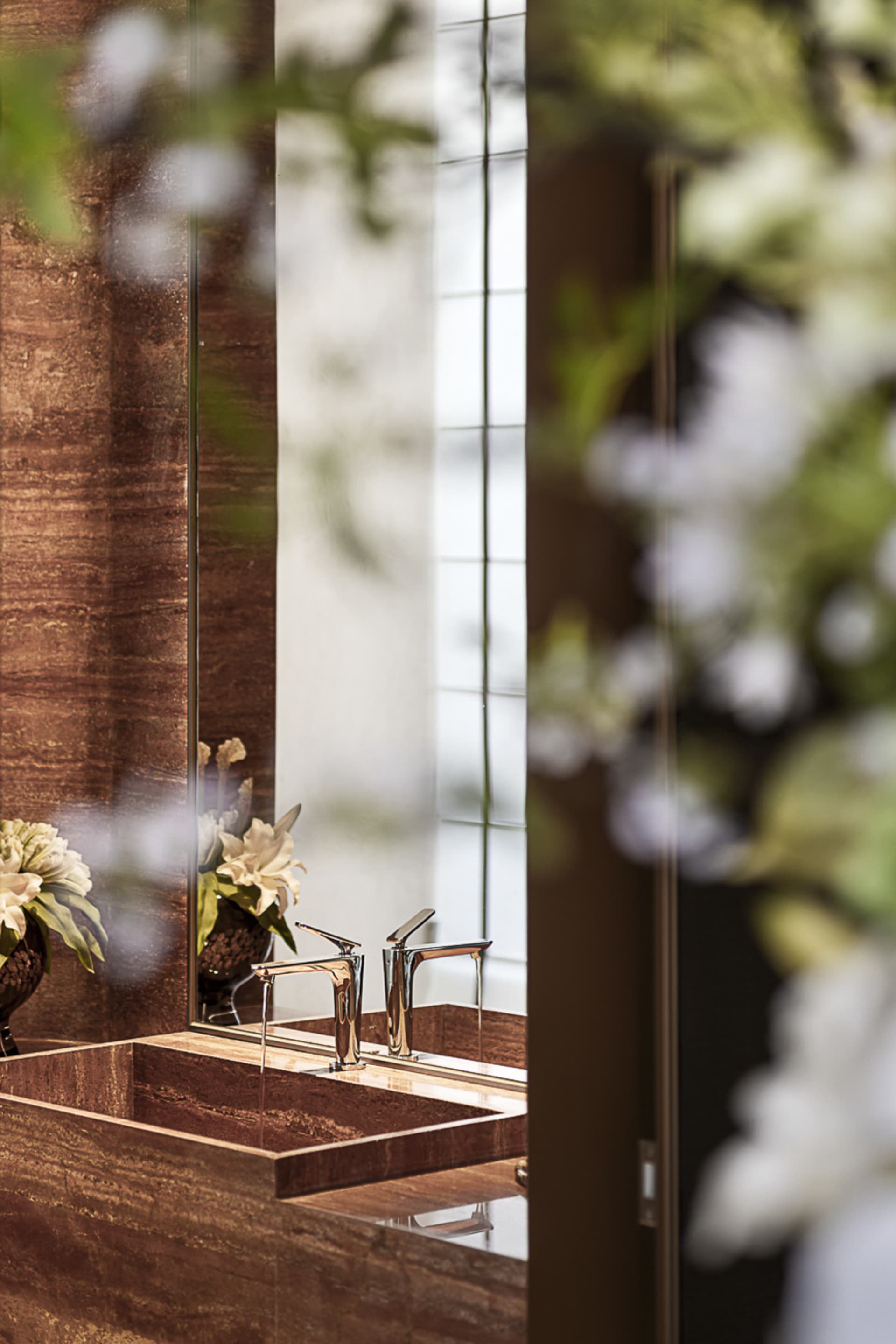 Close-up of a luxurious brown marble bathroom vanity with integrated rectangular sinks and two modern chrome faucets, reflected in a large mirror with white floral arrangements in the foreground.