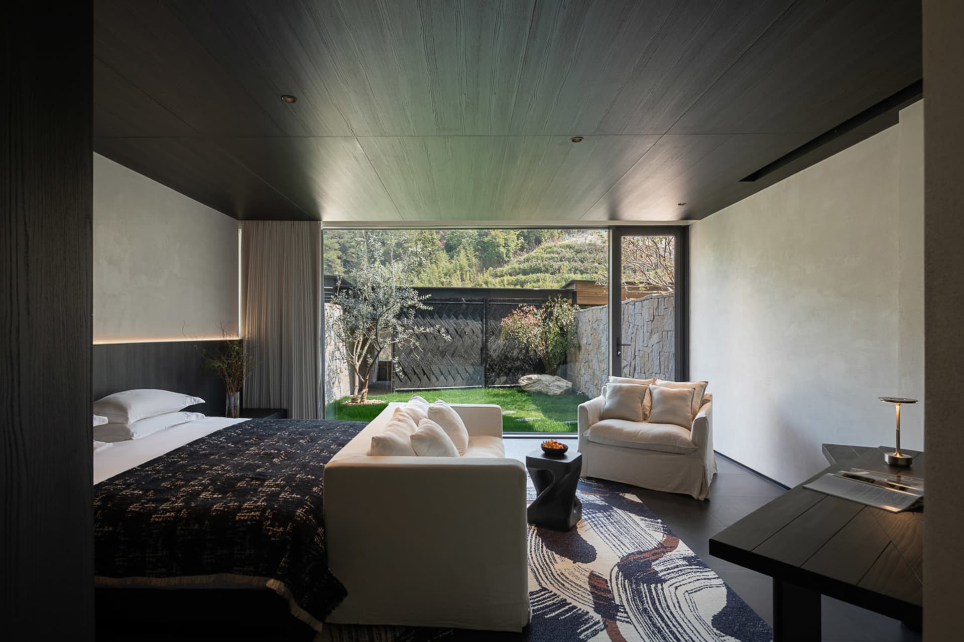 Modern bedroom interior featuring a platform bed with dark patterned bedding, white sofa, patterned area rug, and large glass doors opening to a private green courtyard with trees and stone walls.