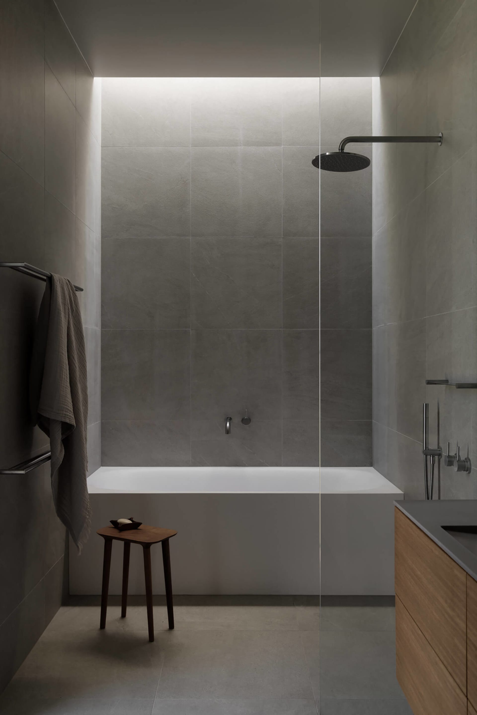 Modern minimalist bathroom featuring large-format concrete tiles, a white freestanding bathtub, black rain shower, wooden vanity, and a small wooden stool with a towel hanging on a black rack.