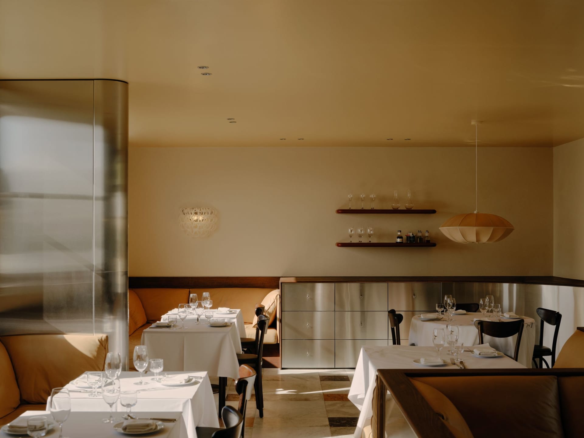 Modern minimalist restaurant interior featuring warm mustard-yellow banquette seating, dark walnut furniture, stainless steel storage cabinet, and large pendant light with white table settings.