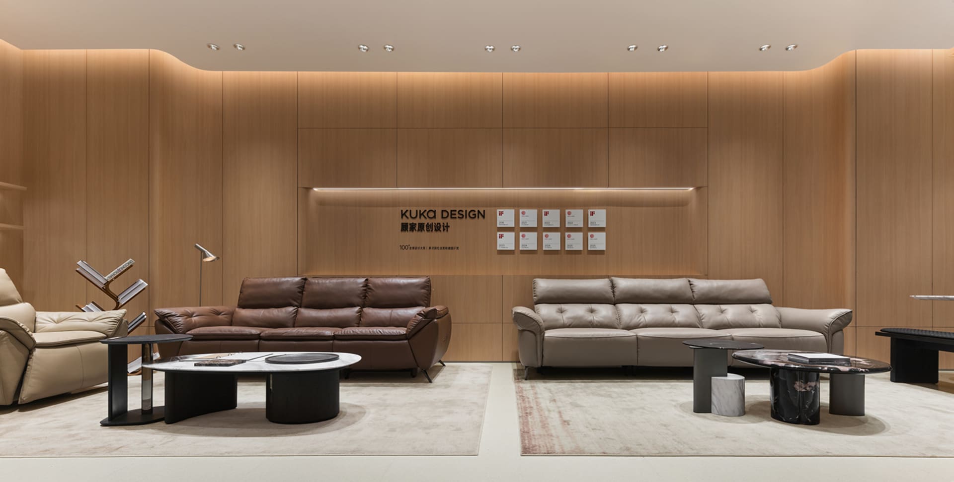 Modern living room display with a brown leather sofa and a light beige leather sofa arranged on separate area rugs in front of a curved wood-paneled wall with recessed lighting and awards.
