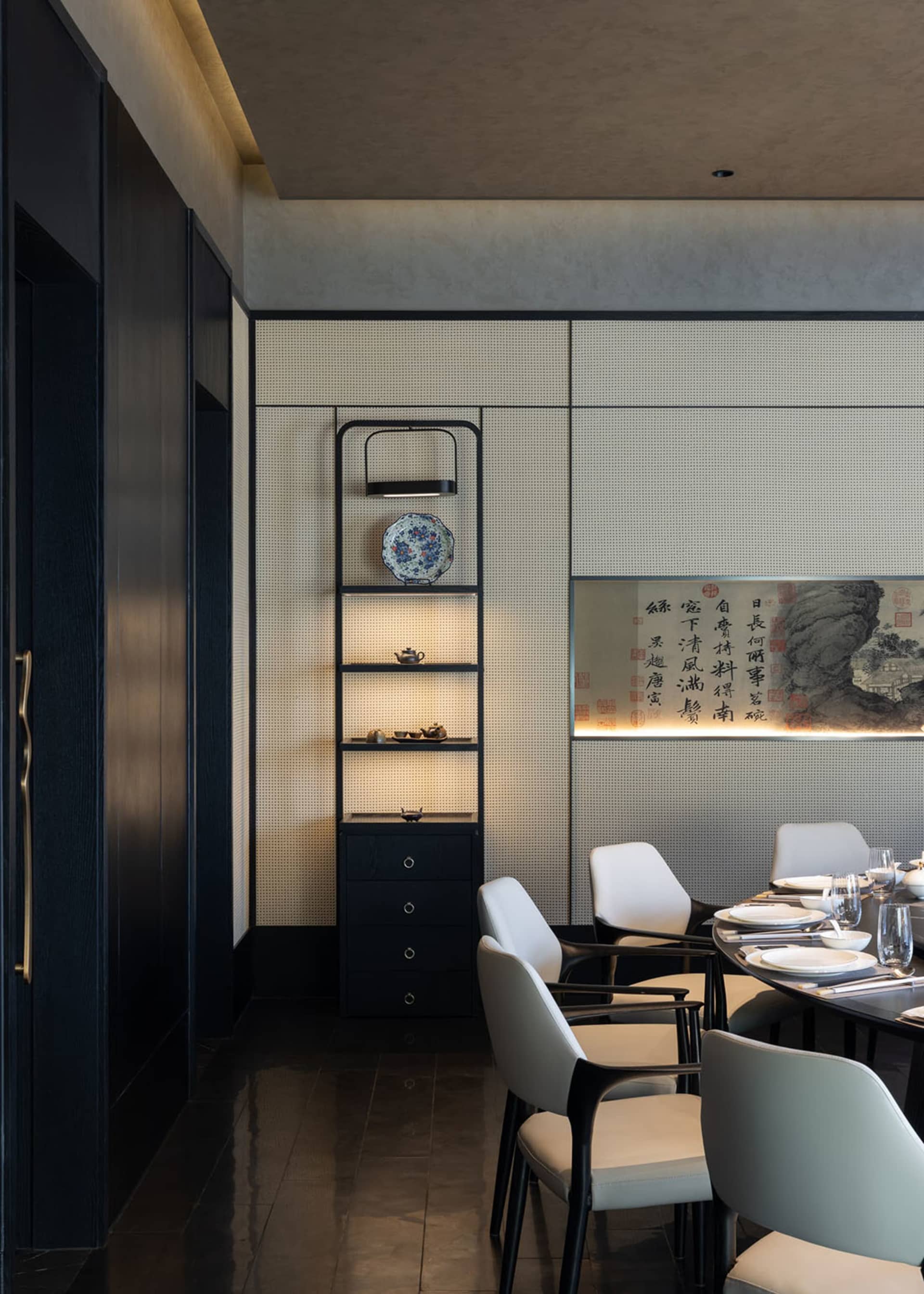 Modern dining room featuring a backlit perforated panel wall, built-in display shelving with blue and white porcelain, a large Chinese calligraphy artwork, and white upholstered dining chairs around a dark table.