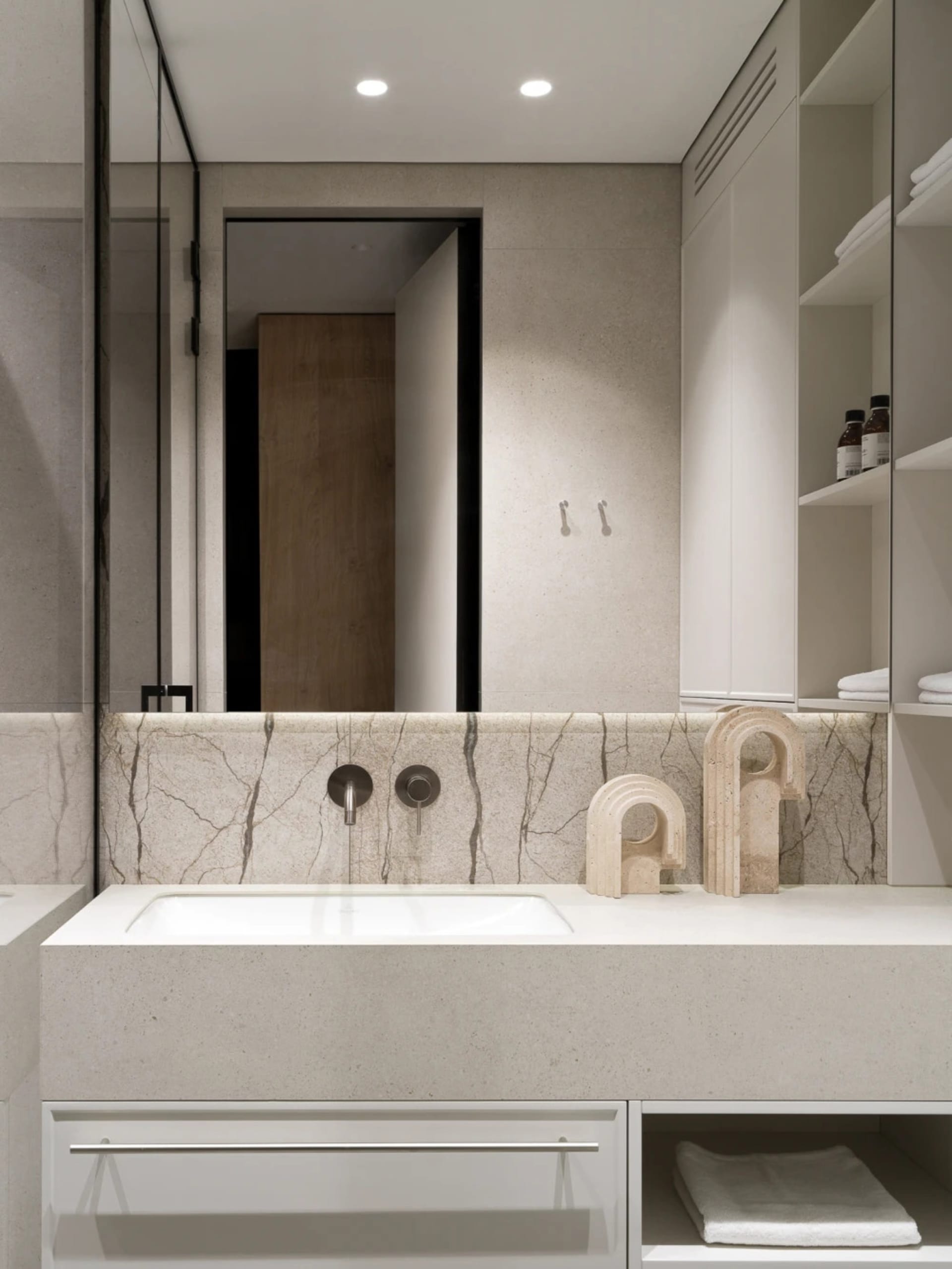 Modern minimalist bathroom vanity with white concrete countertop, integrated sink, marble backsplash, chrome wall-mounted faucet, and decorative travertine sculptures on open shelving.