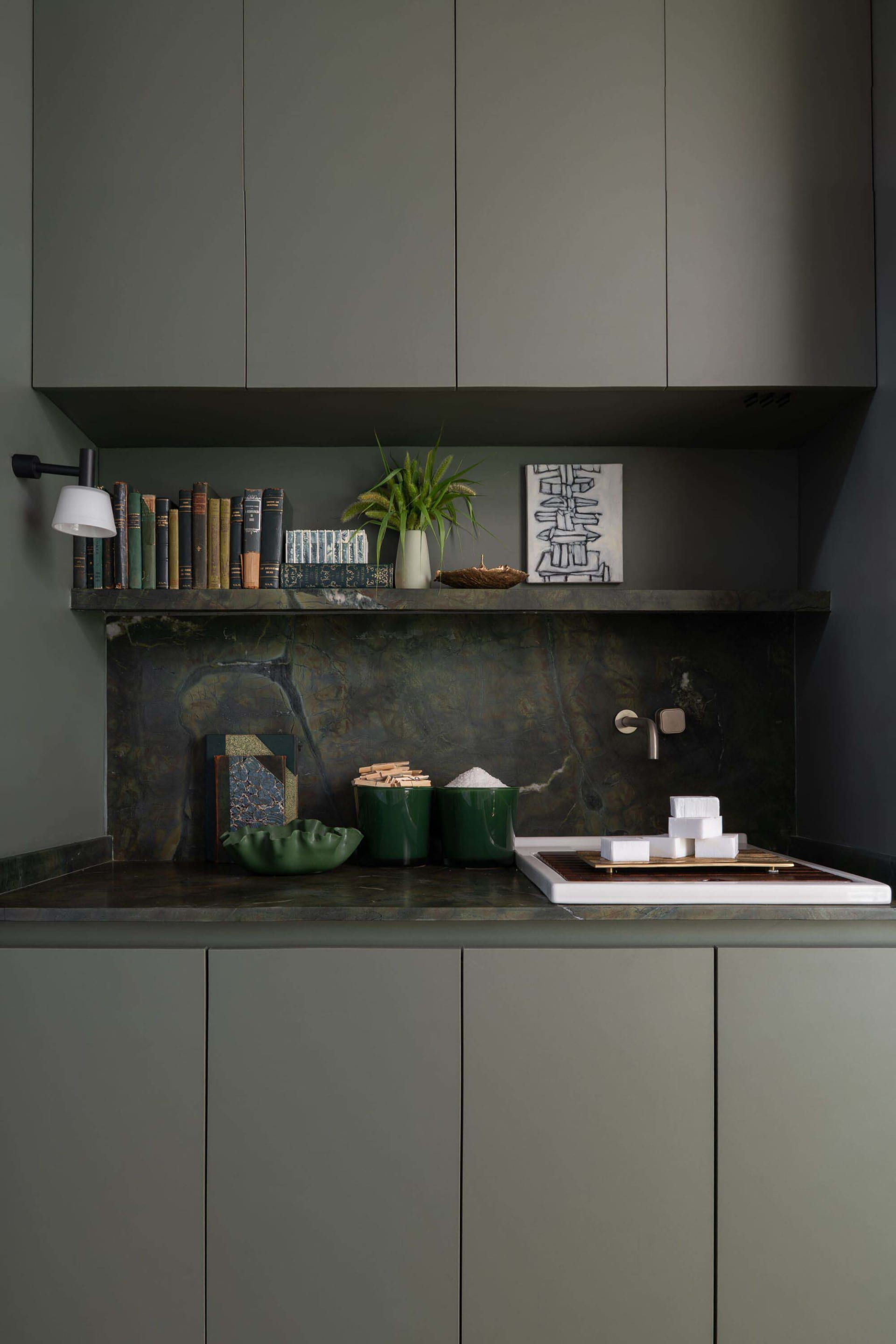 Modern sage green kitchen cabinetry with dark green marble backsplash and countertop, open shelf displaying books, plants, and decor, white ceramic sink with matte faucet.