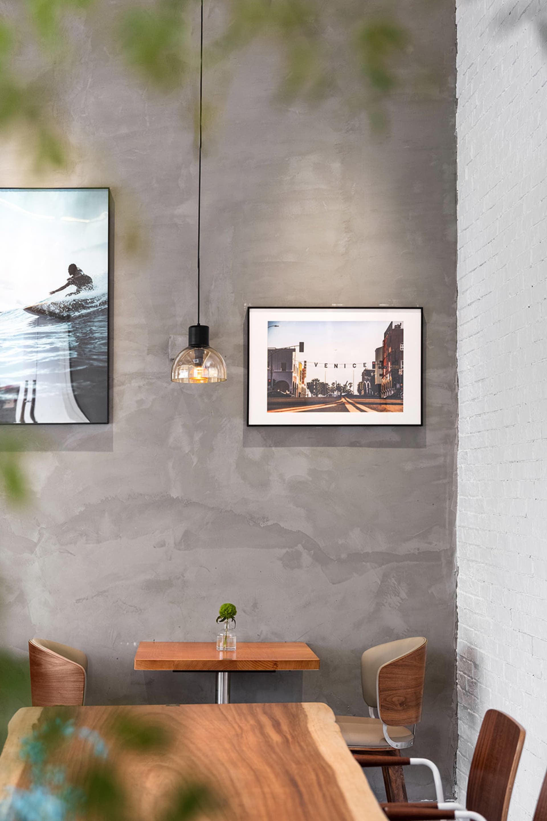 Small wooden dining table against a textured concrete wall with framed surf and Venice Beach photographs, pendant light, and wooden chairs.