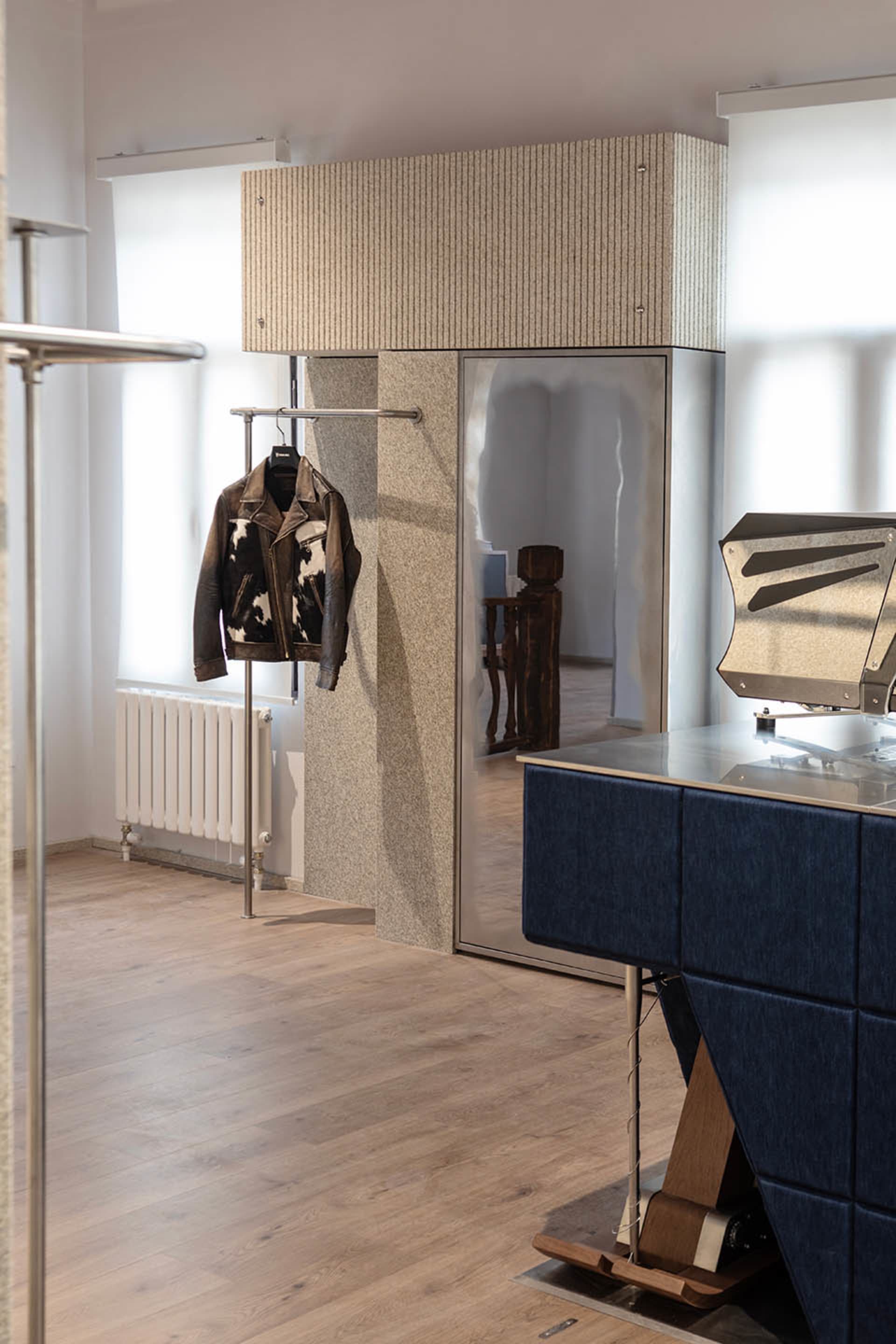 Modern minimalist entryway featuring a tall fluted stone wardrobe with integrated mirror, a blue upholstered console table, and a leather jacket hanging on a chrome rack against a light wood floor.
