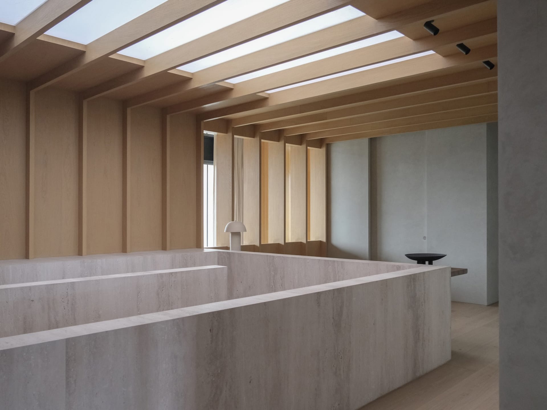 Modern minimalist living room featuring massive travertine blocks forming sunken seating, light wood ceiling beams, and long rectangular skylights allowing natural daylight.