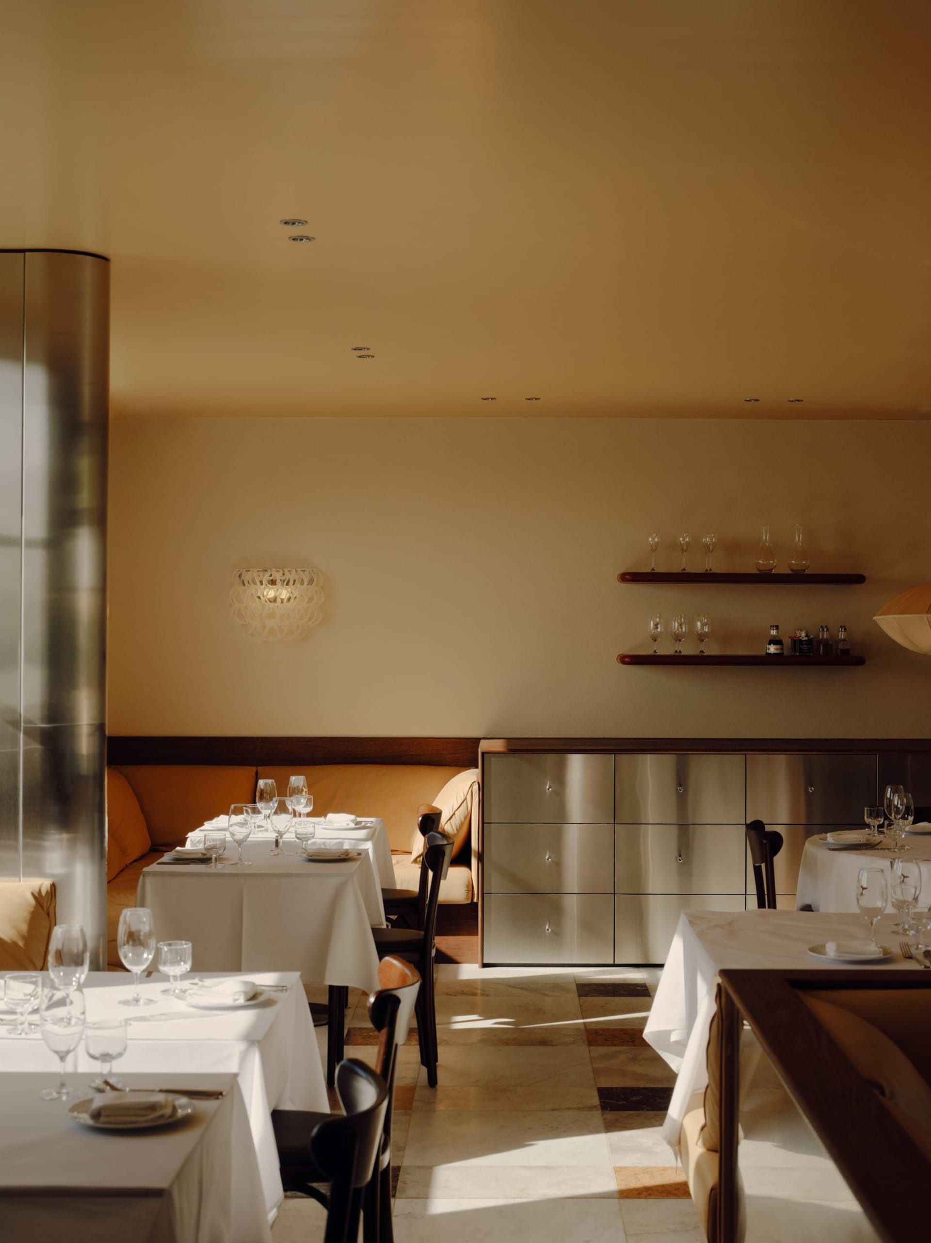 Elegant fine dining restaurant interior with white tablecloths, dark wood chairs, orange banquette seating, stainless steel storage cabinet, and sculptural wall sconce under a high ceiling.