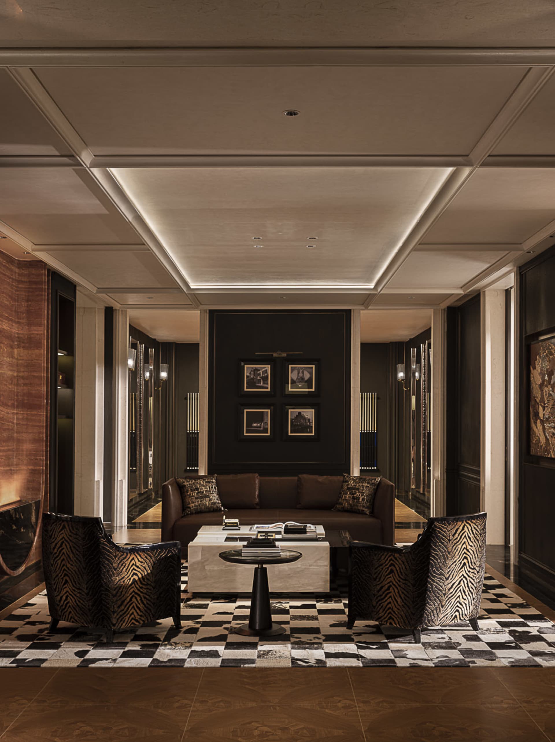 Luxurious hotel lobby lounge with two zebra-print velvet armchairs, a brown leather sofa, white marble coffee table, and black-and-white checkerboard cowhide rug under dramatic coffered ceiling with cove lighting.
