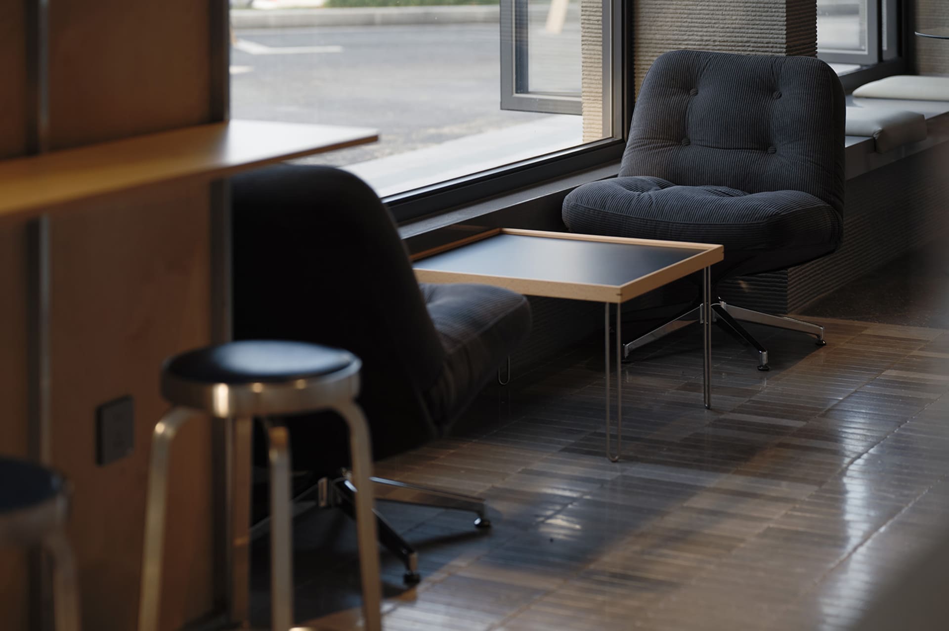 Modern corduroy lounge chair with chrome swivel base placed next to a large window with a wooden side table and black stool in foreground