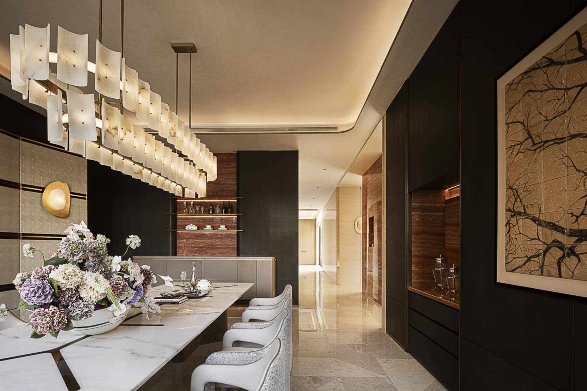 Modern luxury dining room featuring a long white marble dining table, six light grey upholstered dining chairs, a dramatic multi-tiered alabaster chandelier, dark wood paneling, and a large framed ink painting of tree branches.