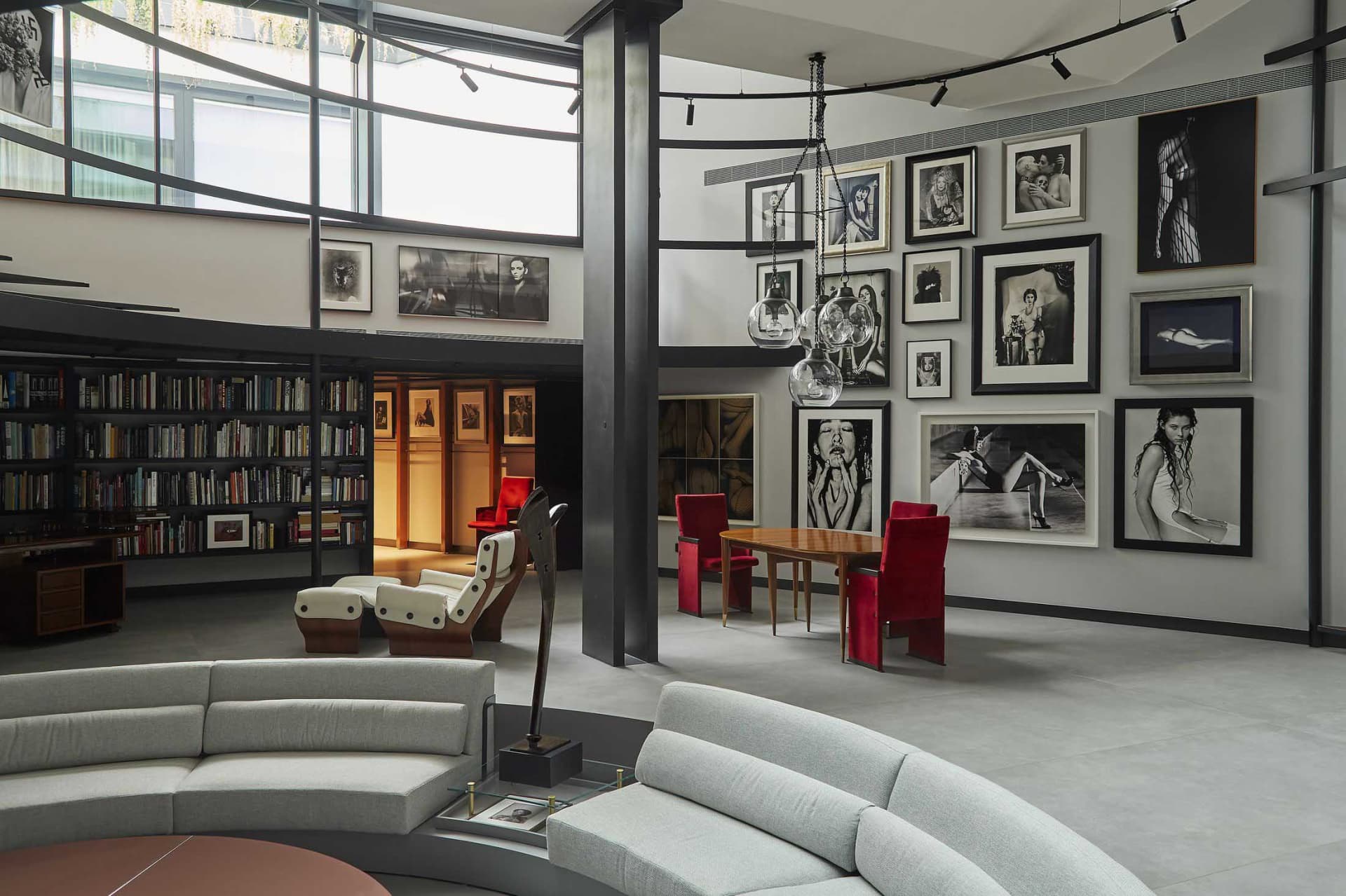 Modern double-height living room with curved light-grey sectional sofas, a large gallery wall of black and white portrait photography, red velvet dining chairs, wooden table, and industrial metal structural columns under a skylight.
