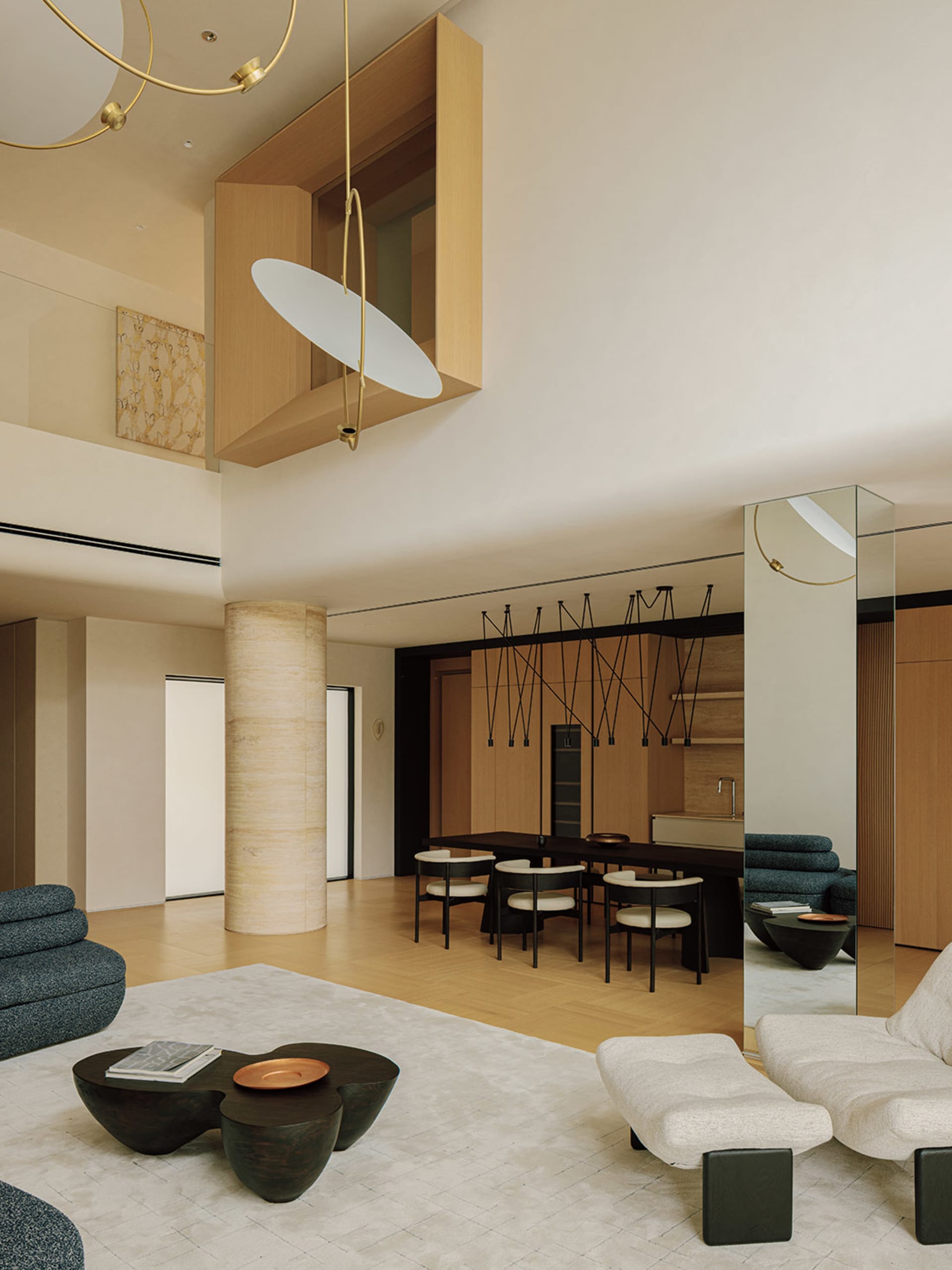 Modern double-height living room featuring a large cylindrical travertine column, light wood architectural volumes, sculptural brass and white pendant light, dark dining table with white chairs, and contemporary low seating in blue and cream tones.