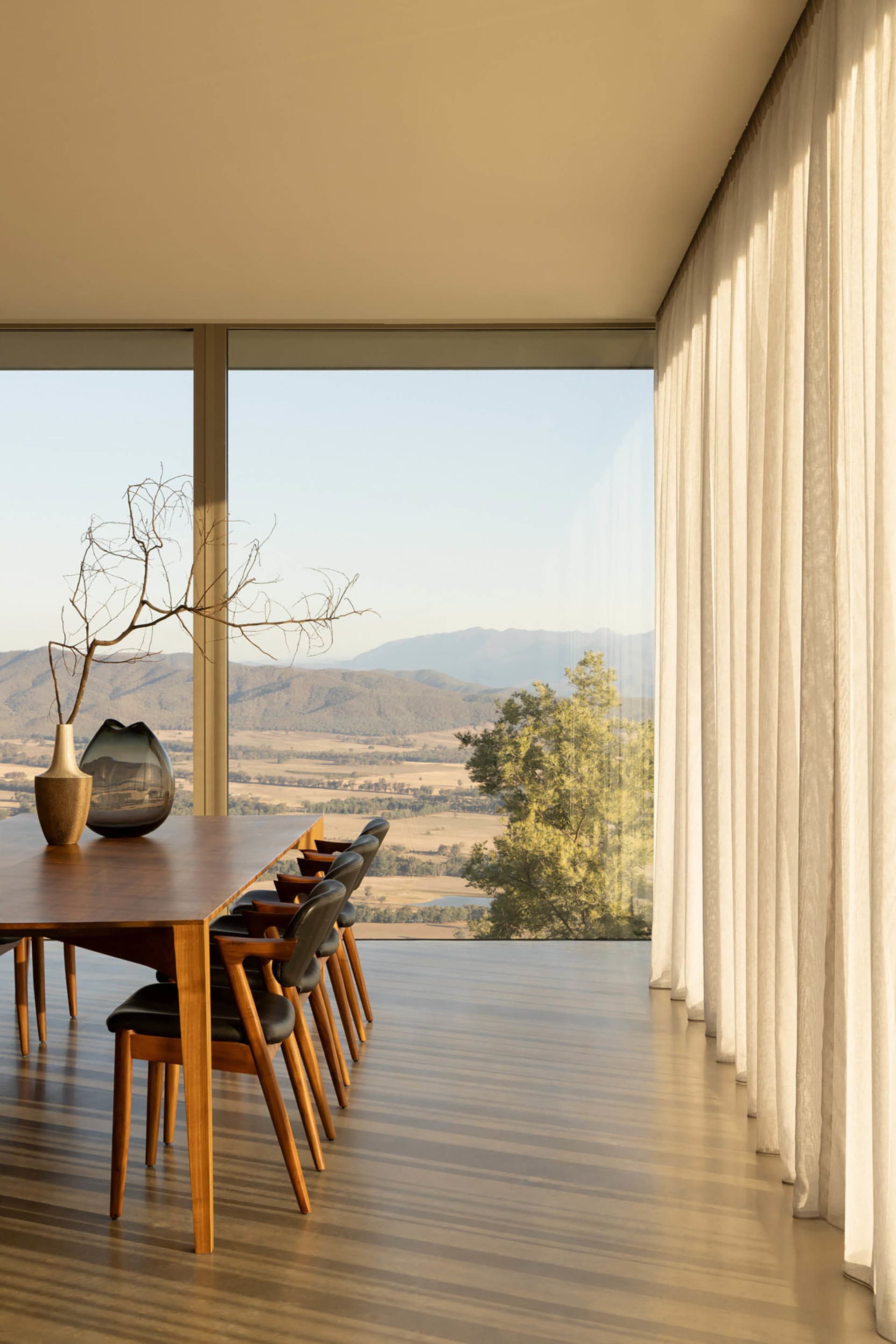 Modern dining room featuring a long walnut table, black leather dining chairs, large floor-to-ceiling windows overlooking mountains and fields, with sheer white curtains and minimalist decor.